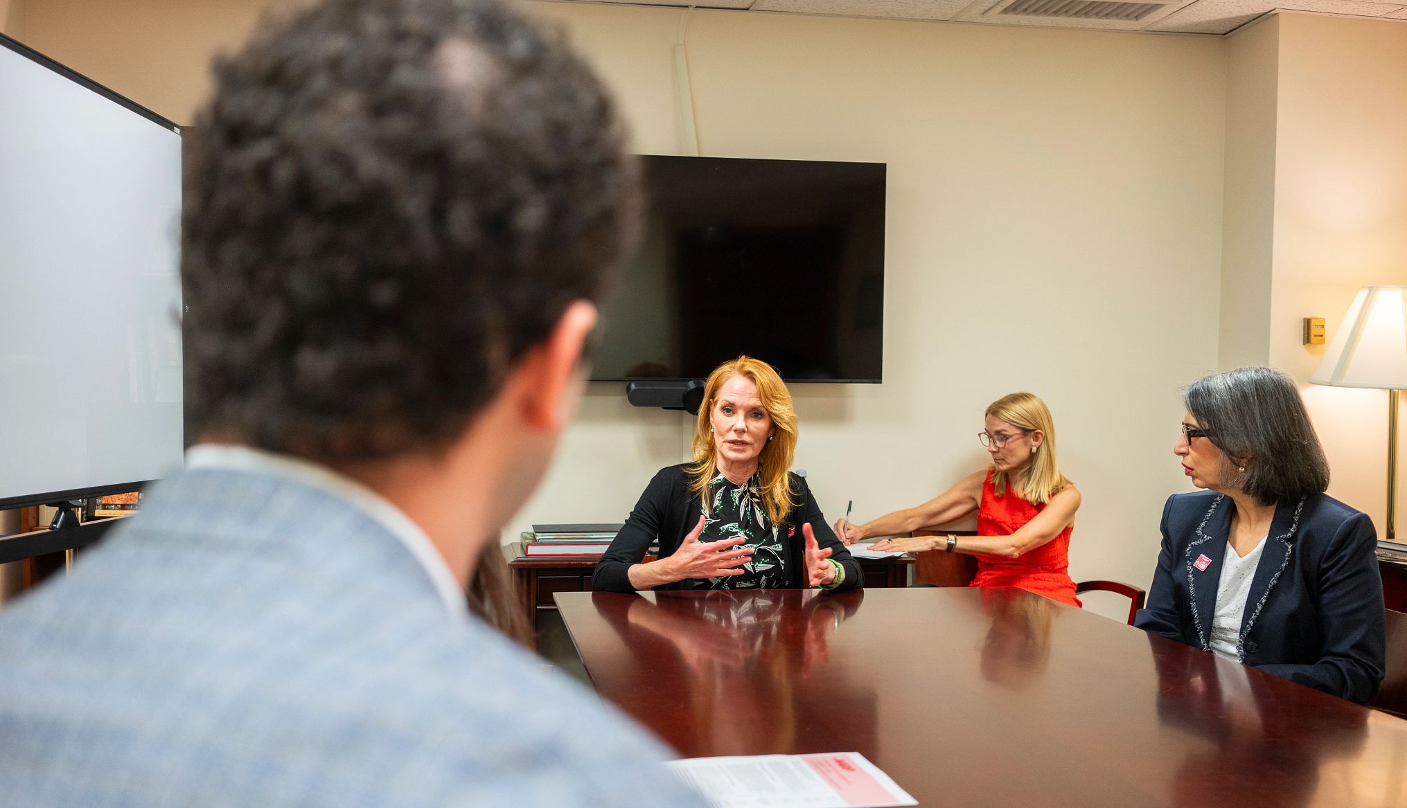 Marg Helgenberger Marg Helgenberger, in a printed black dress and black sweater, speaks while sitting at a conference table.