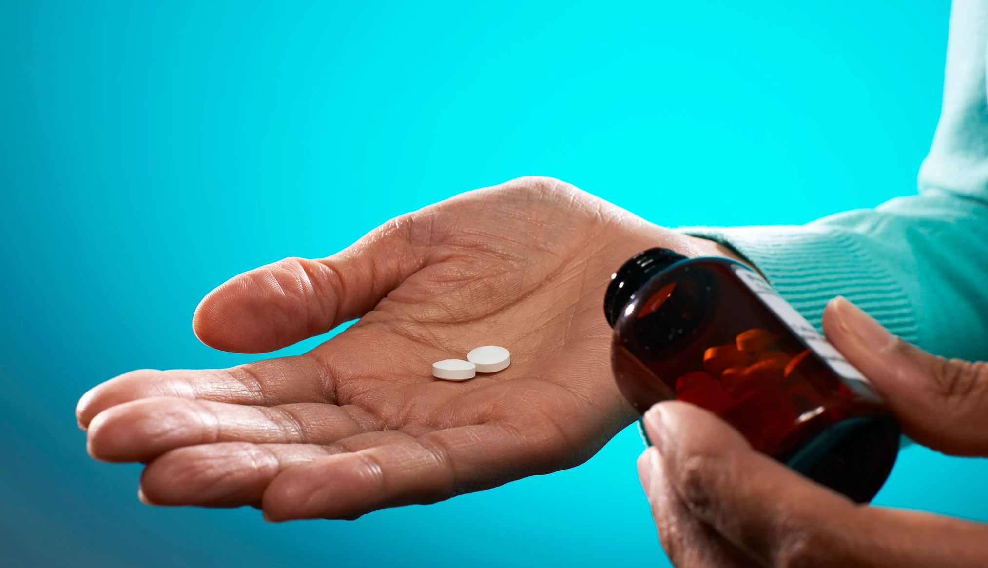 AARP fights to lower prescription drug costs Close up of person holding tablets and brown pill bottle, on blue background.