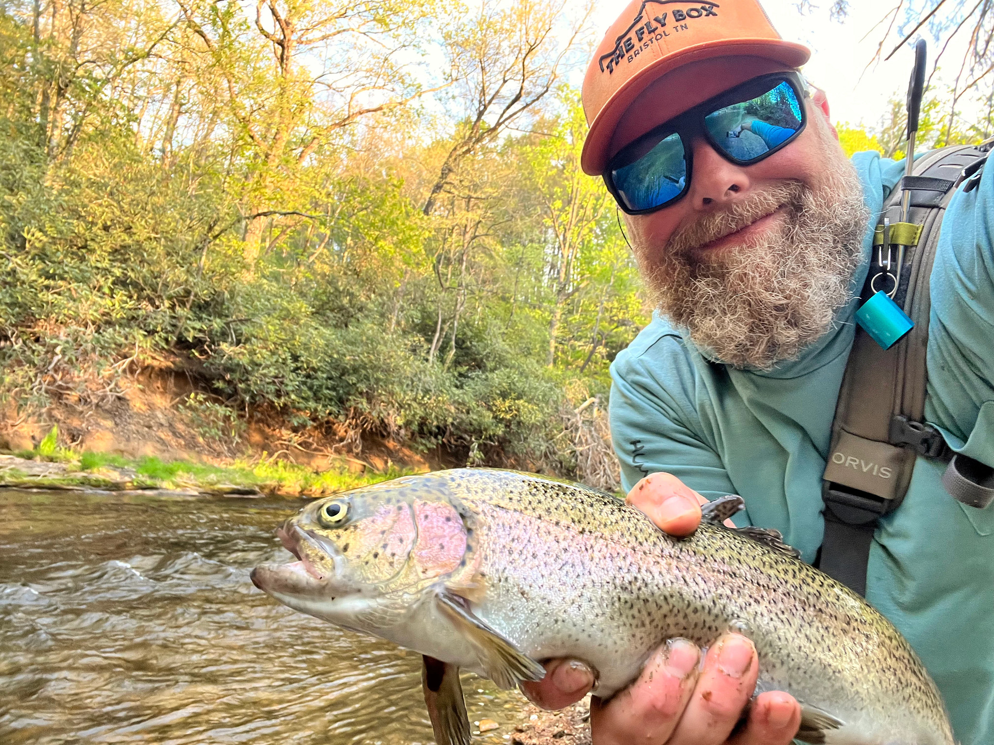 A selfie of Robert Annis holding a large caught rainbow trout in his hands while standing in front of a flowing river lined with green trees.