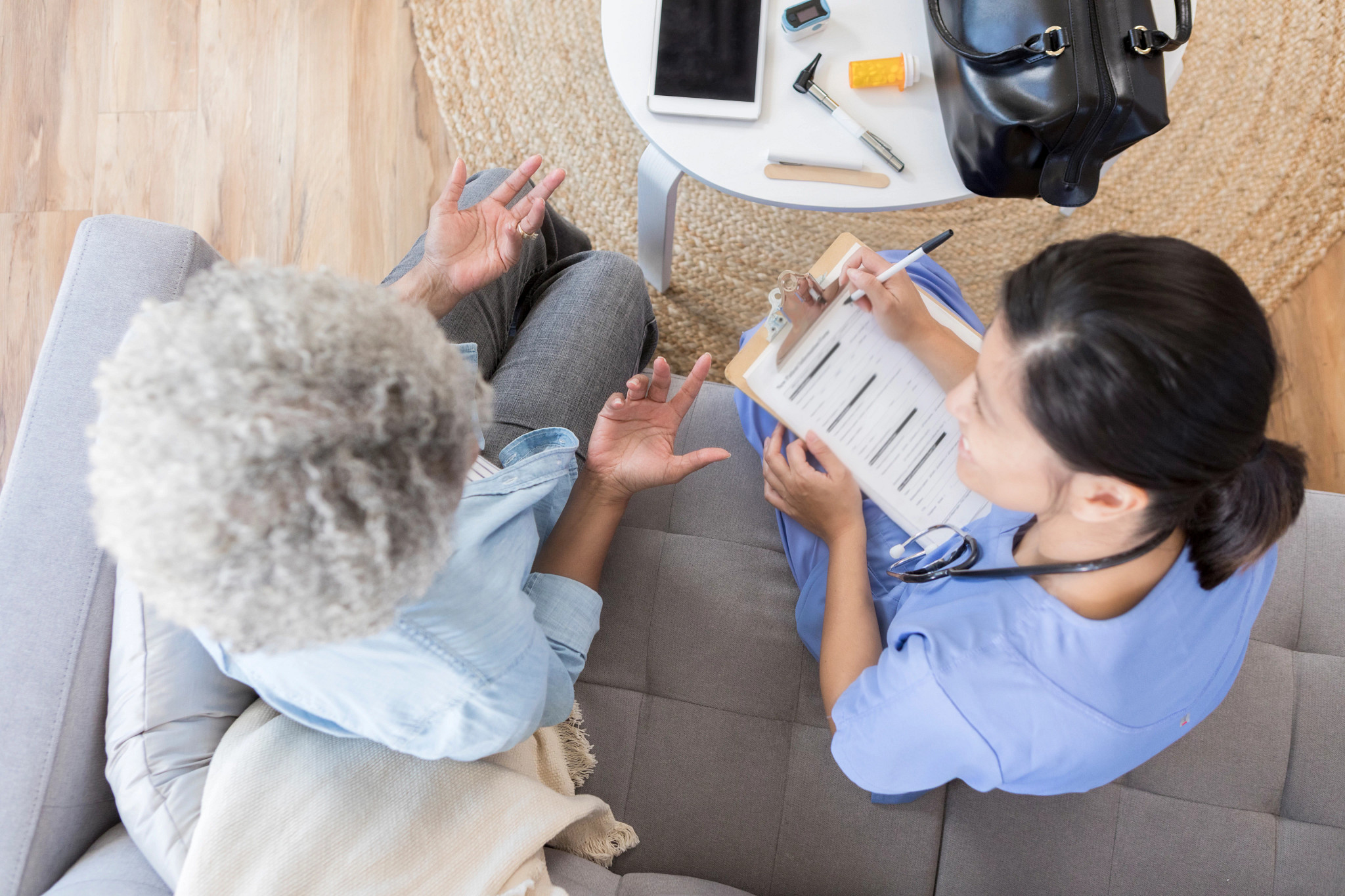 In this bird's eye view, a female home health nurse wearing scrubs sits on a living room couch next to a senior female patient.  She takes notes as she listens to her patient.  There is medical equipment and a digital tablet on the coffee table.