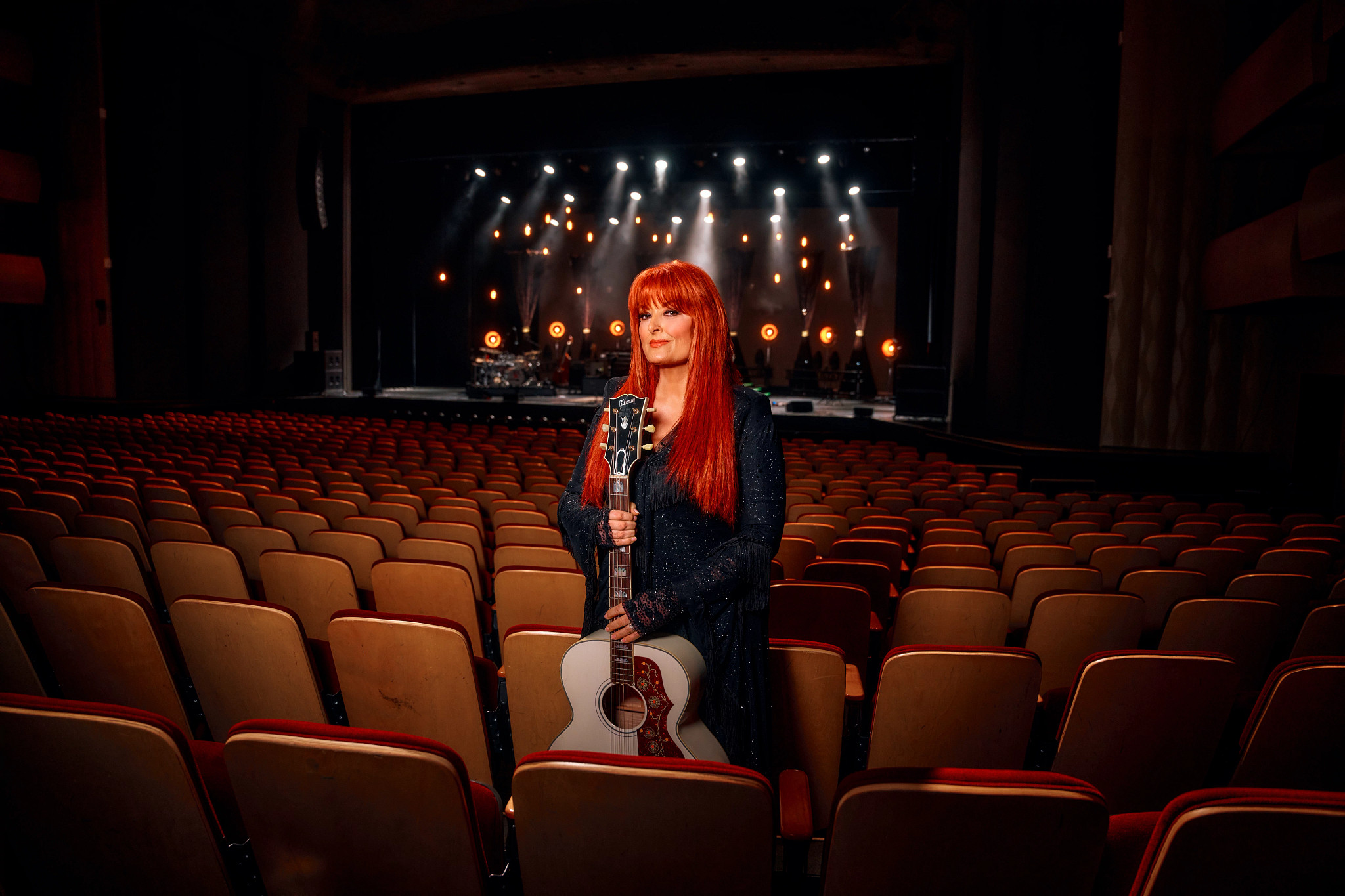 Wynonna Judd posing with a guitar in an auditorium.