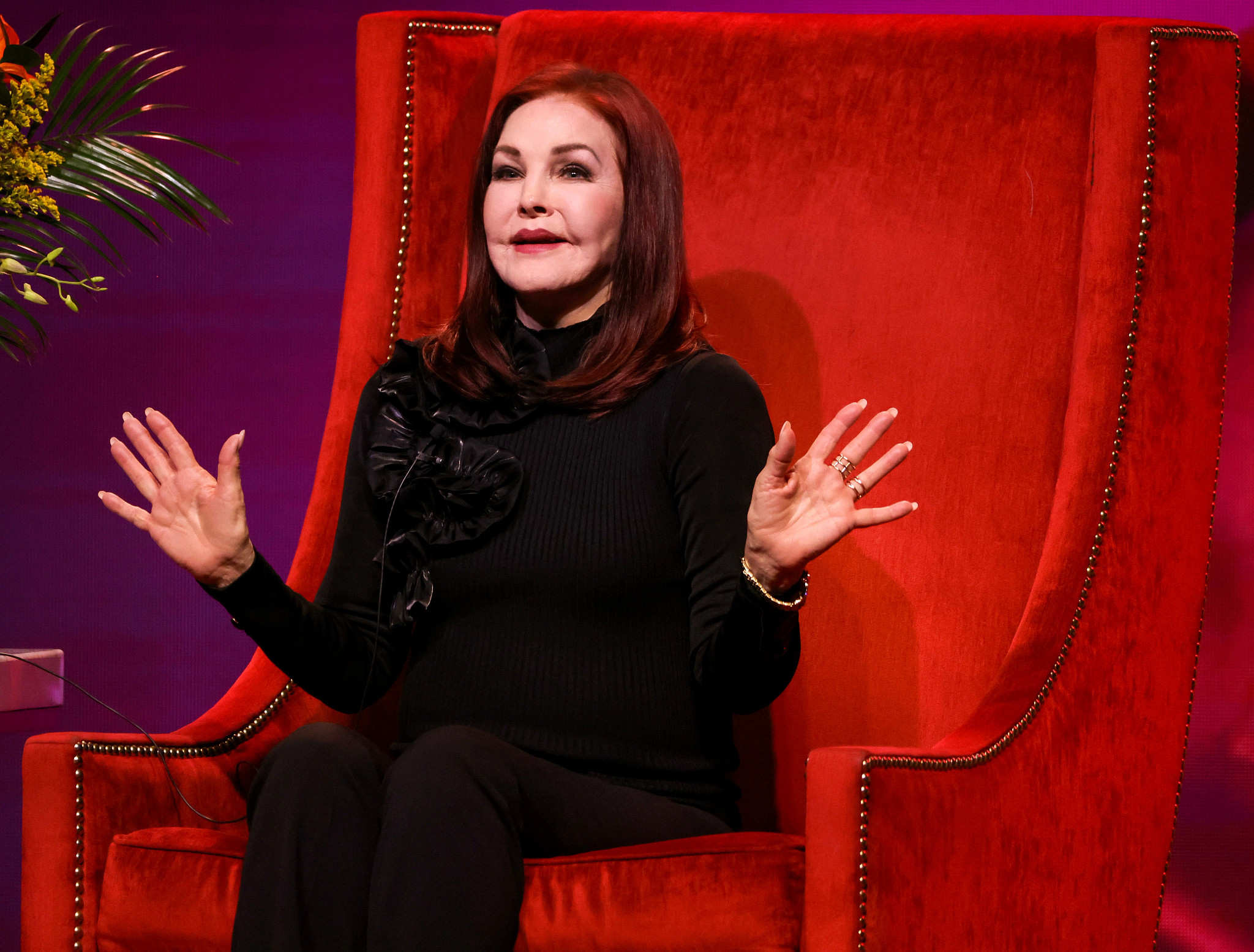 priscilla presley emoting while speaking, seated in a red upholstered chair