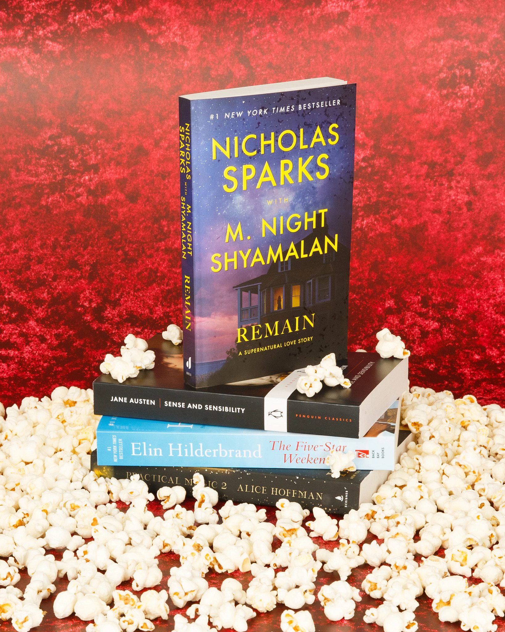 A colorful book display features “Remain” by Nicholas Sparks and M. Night Shyamalan standing upright atop a stack of novels, surrounded by scattered popcorn against a bold red backdrop