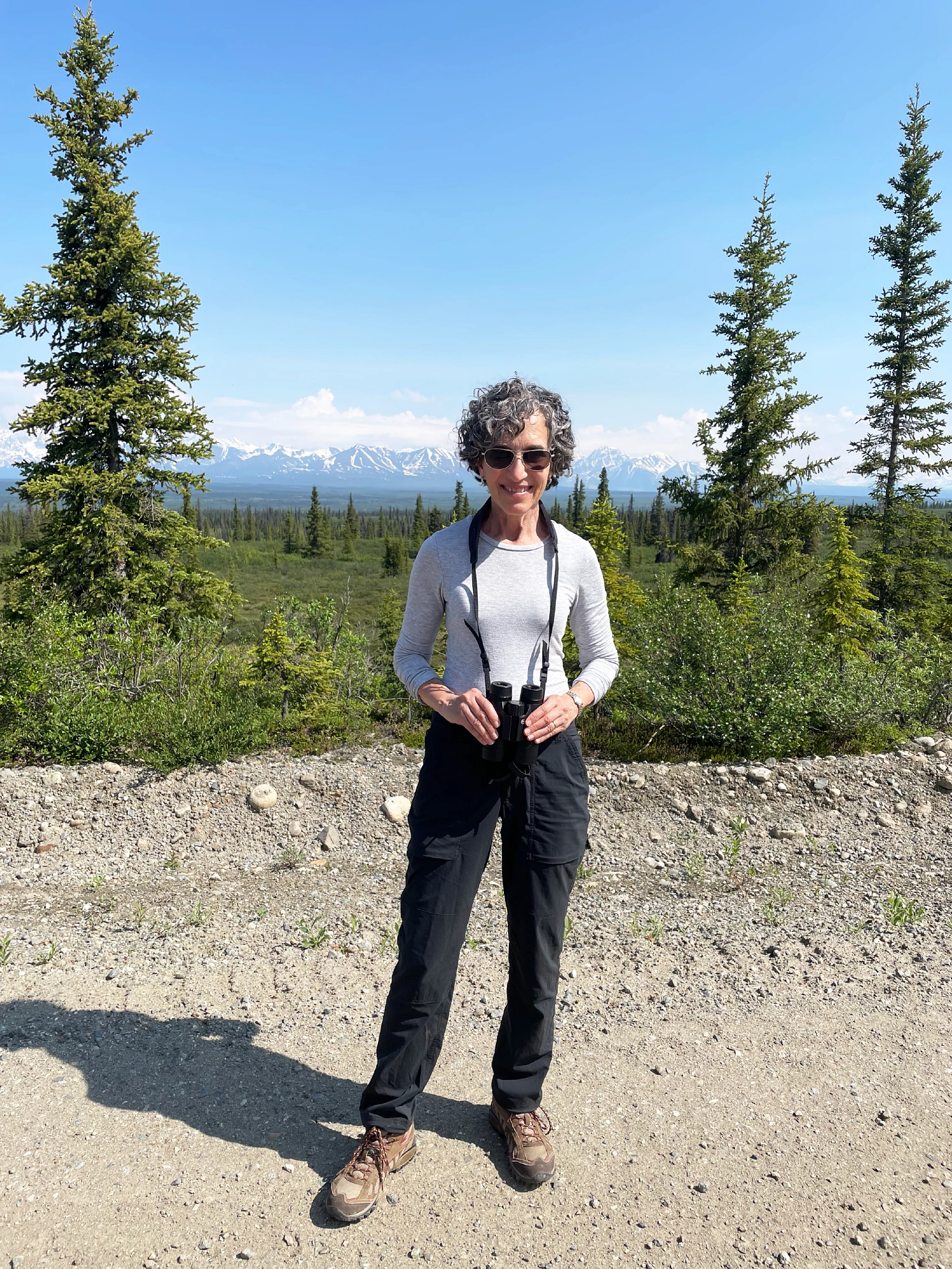 a woman holding binoculars with trees and mountains behind her