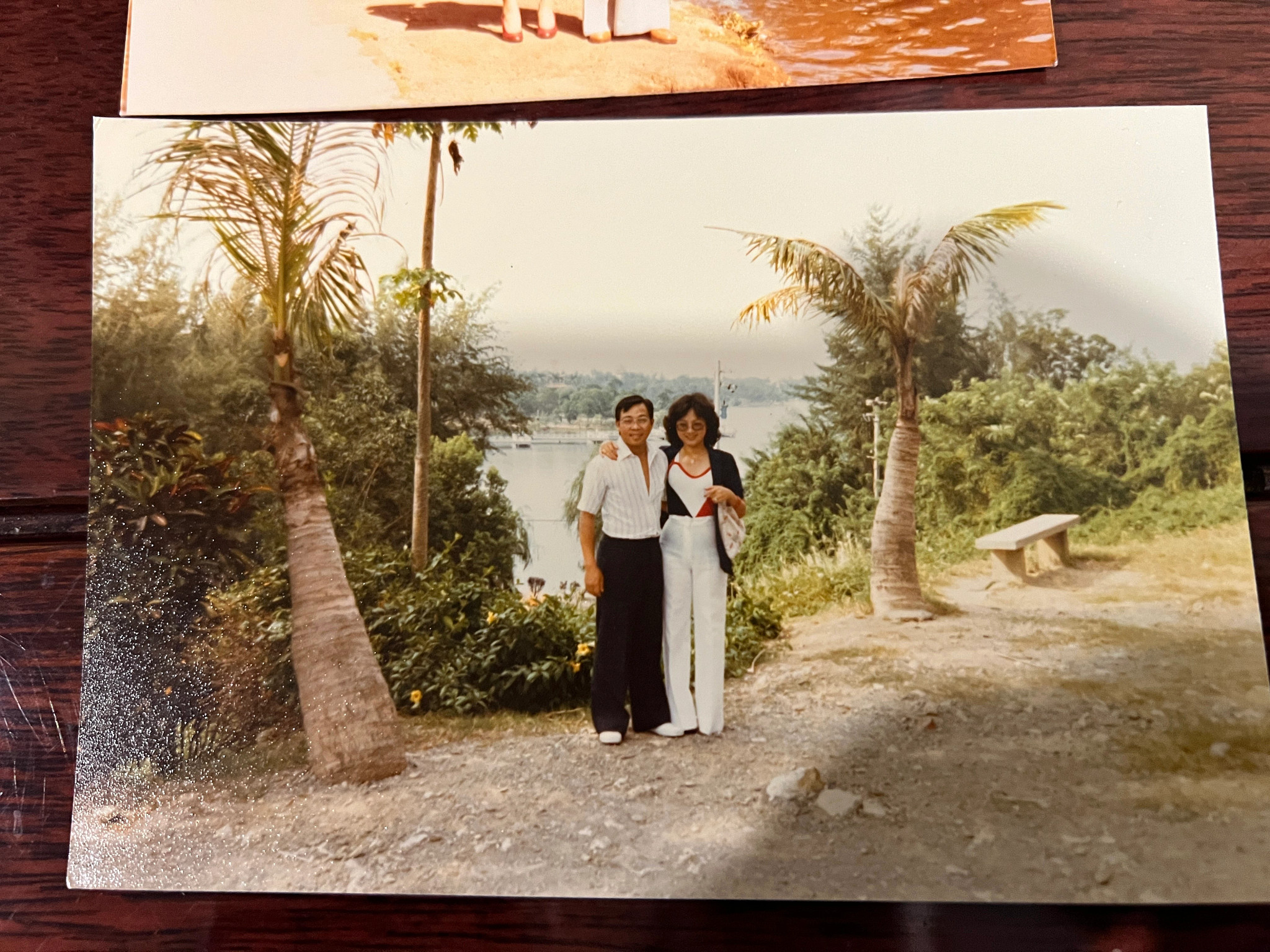 a man and woman pose on a trail. trees and bushes are behind them, with water in the distance