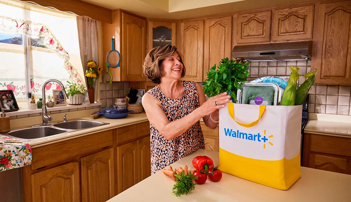 Smiling woman places fresh groceries into a Walmart+ bag on her kitchen counter.