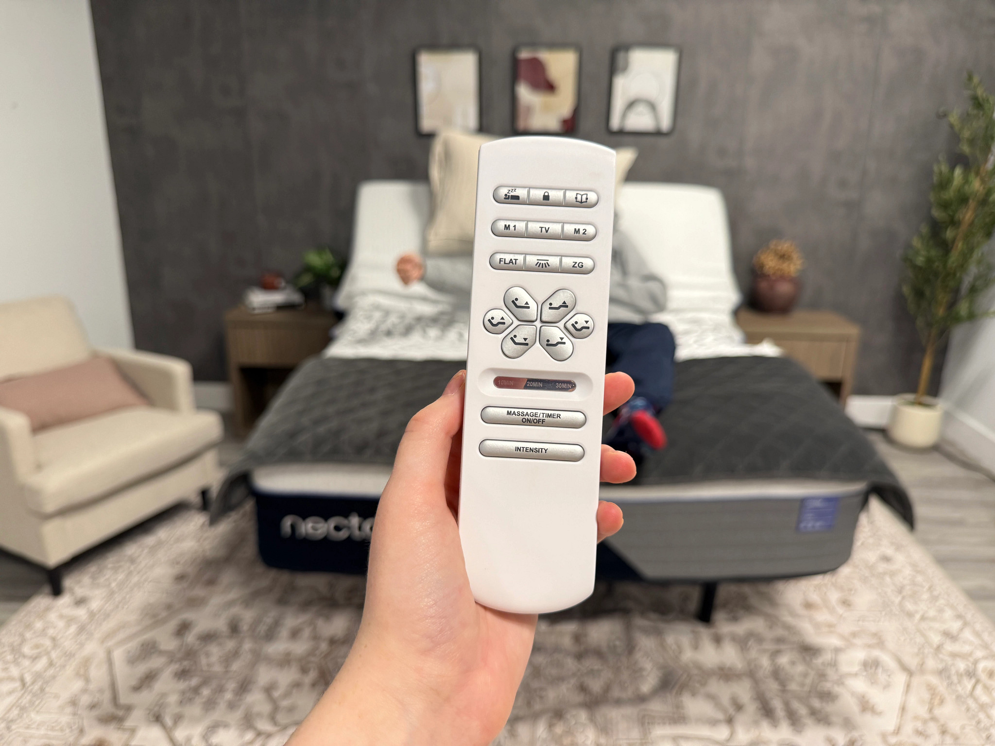 The remote control for a Nectar Premier Adjustable Base is held in the foreground while in the background a man lies on the base.