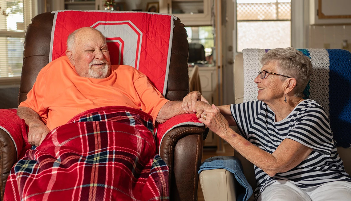 A disabled senior Caucasian male and his wife hold hands while sitting in their living room