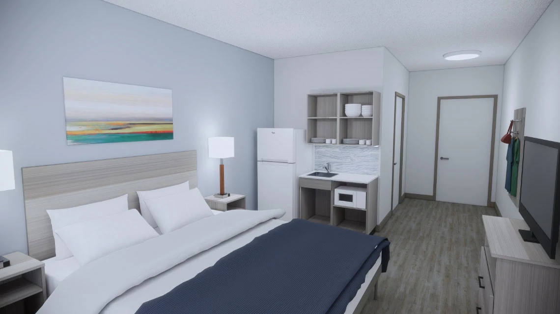 Suburban Studios® bed in a hotel room with white and blue linens against a light gray wall with a refrigerator, small kitchen, TV, hardwood floors