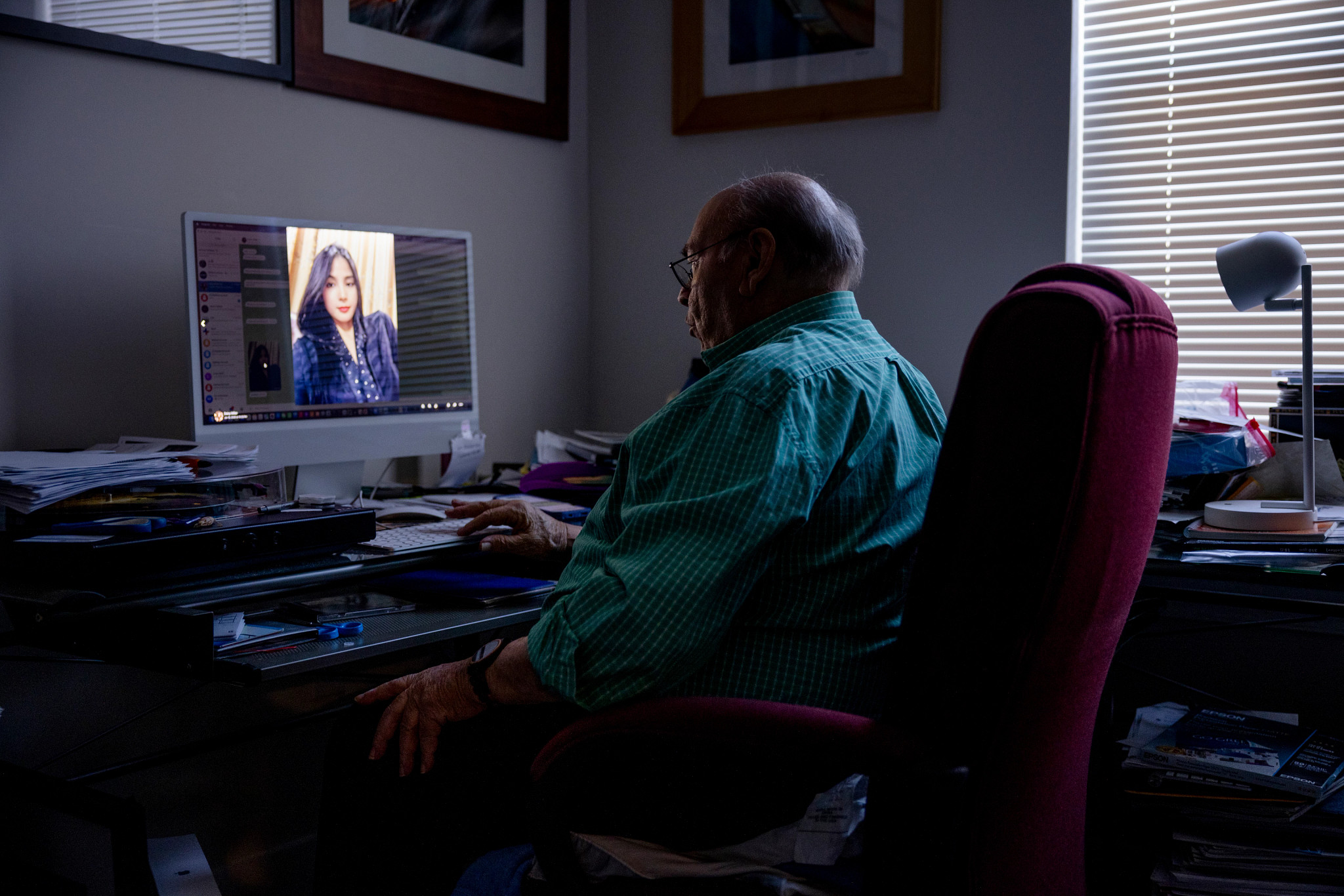 a man sits at a desk in a darkened room. he is looking at a computer screen with a woman's photo on it.