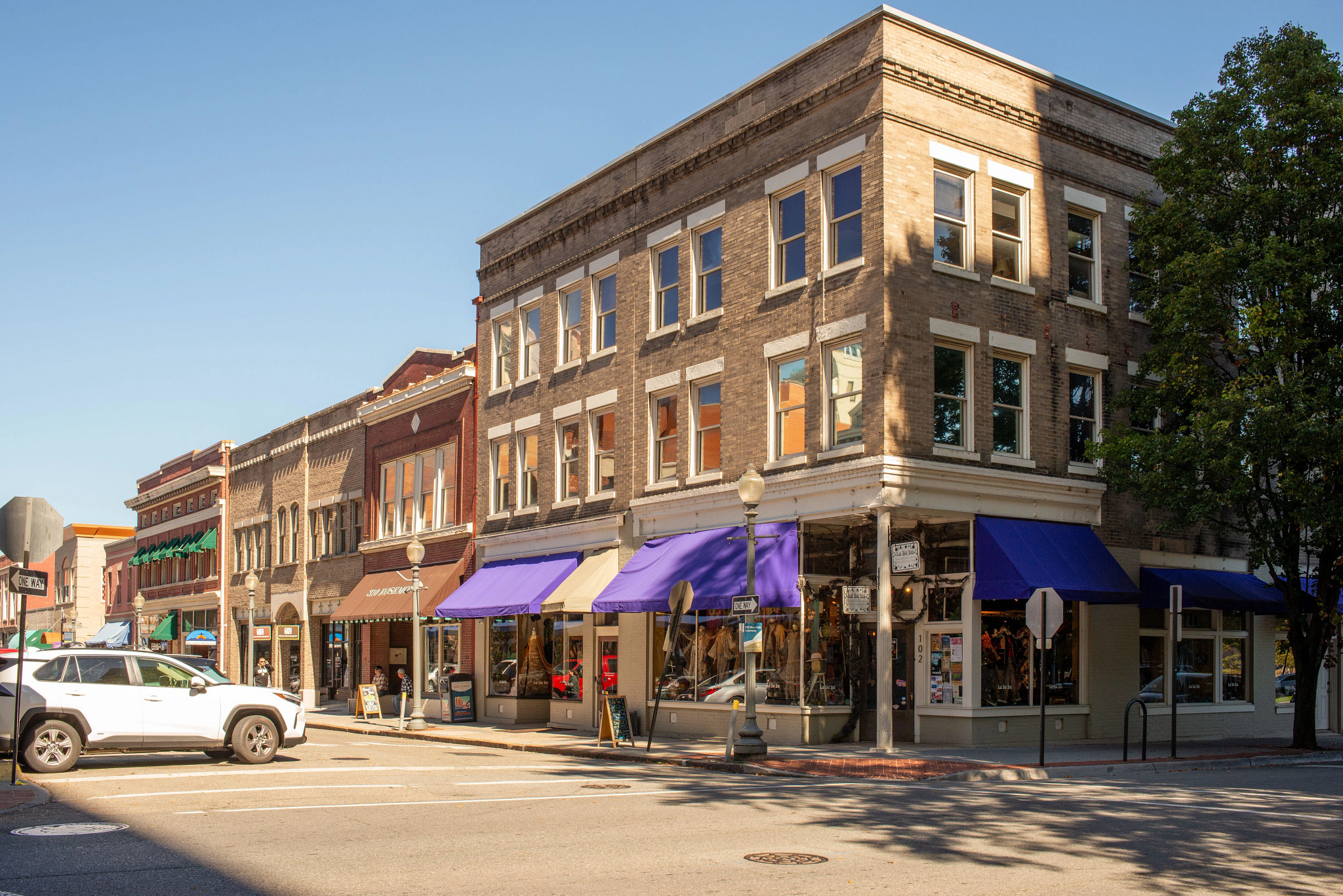 Historic downtown street corner featuring a multi-story brick commercial building with colorful storefront awnings, local shops, walkable sidewalks, and a vibrant small-business district