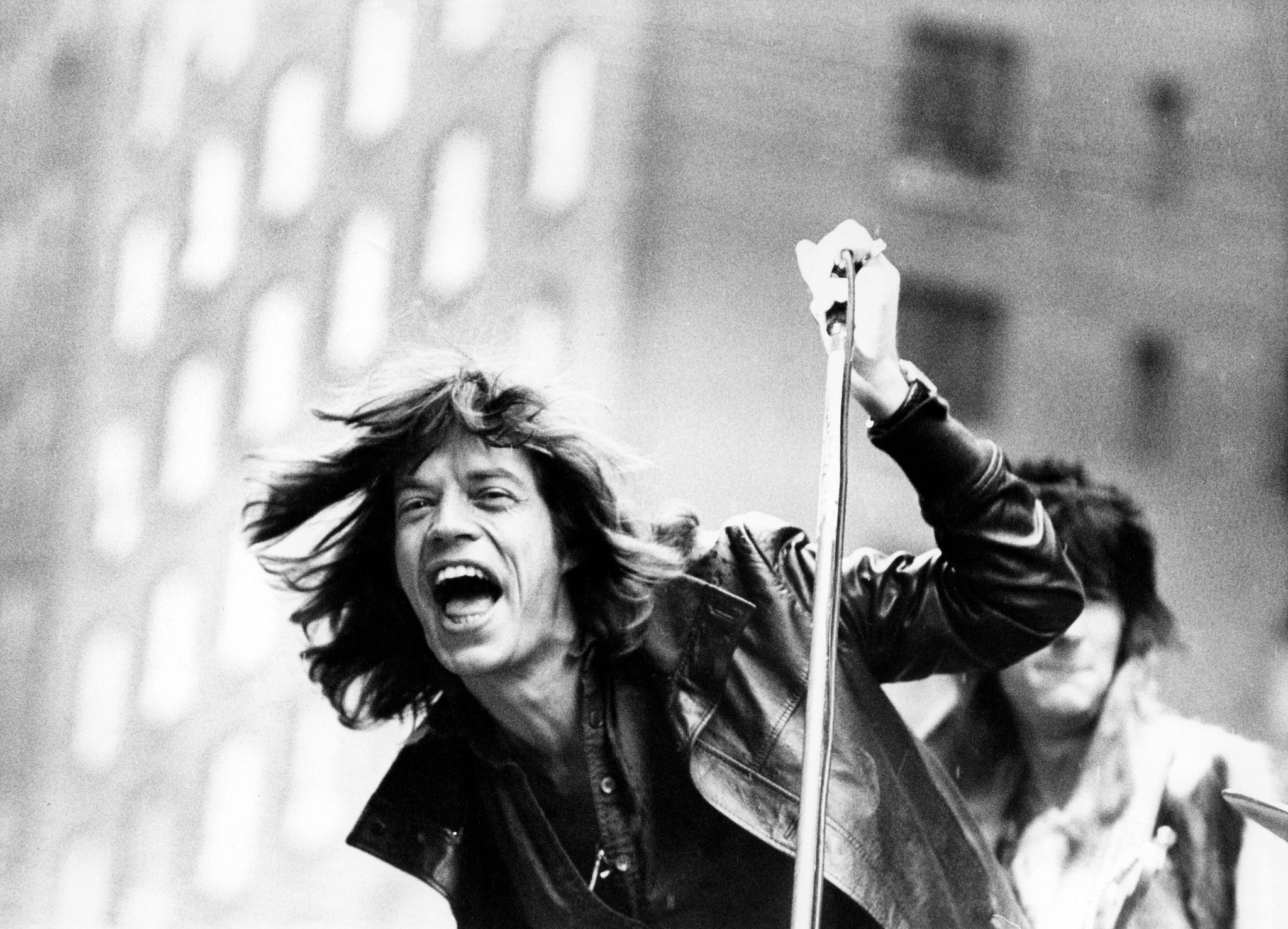 A black-and-white archival photograph of a young Mick Jagger singing passionately into a microphone during a live outdoor performance. He has long, wind-blown hair, wears a leather jacket, and stands in front of guitarist Ronnie Wood, who is partially visible in the background.