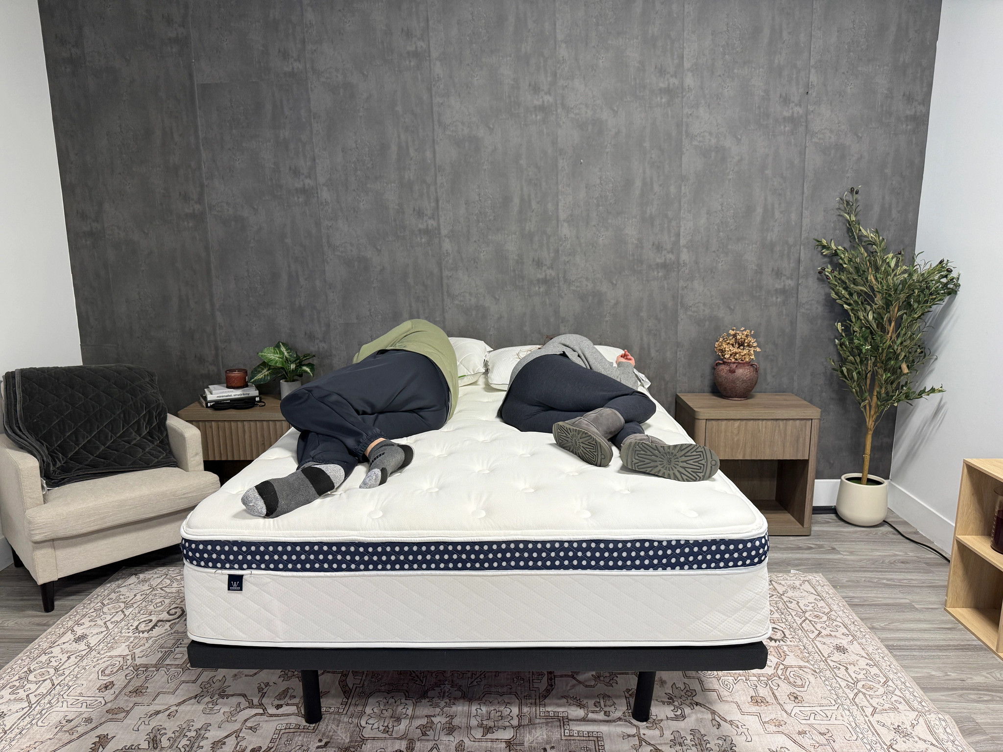 Two people lie on a mattress in a room with a plant, chair and beside table