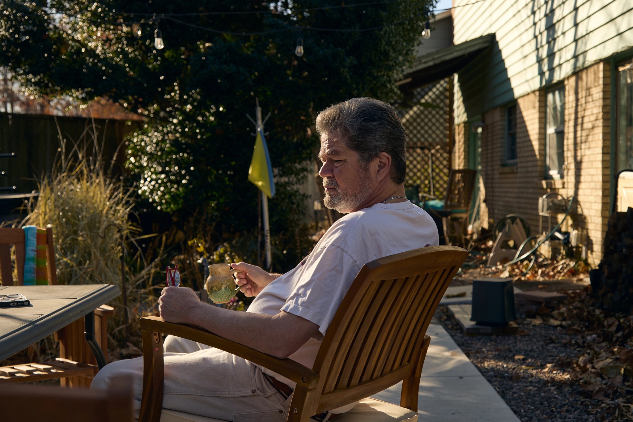 a man, holding a mug, sits in an outdoor chair in the yard of his home
