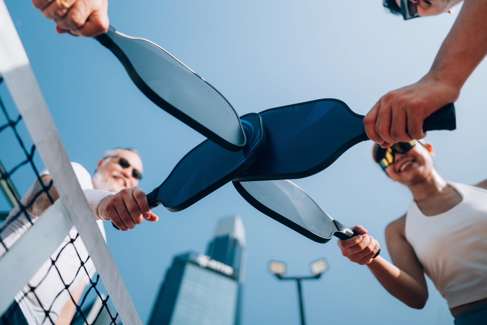 A low-angle view shows a group of four adults tapping paddles to show sportsmanship after a pickleball game.