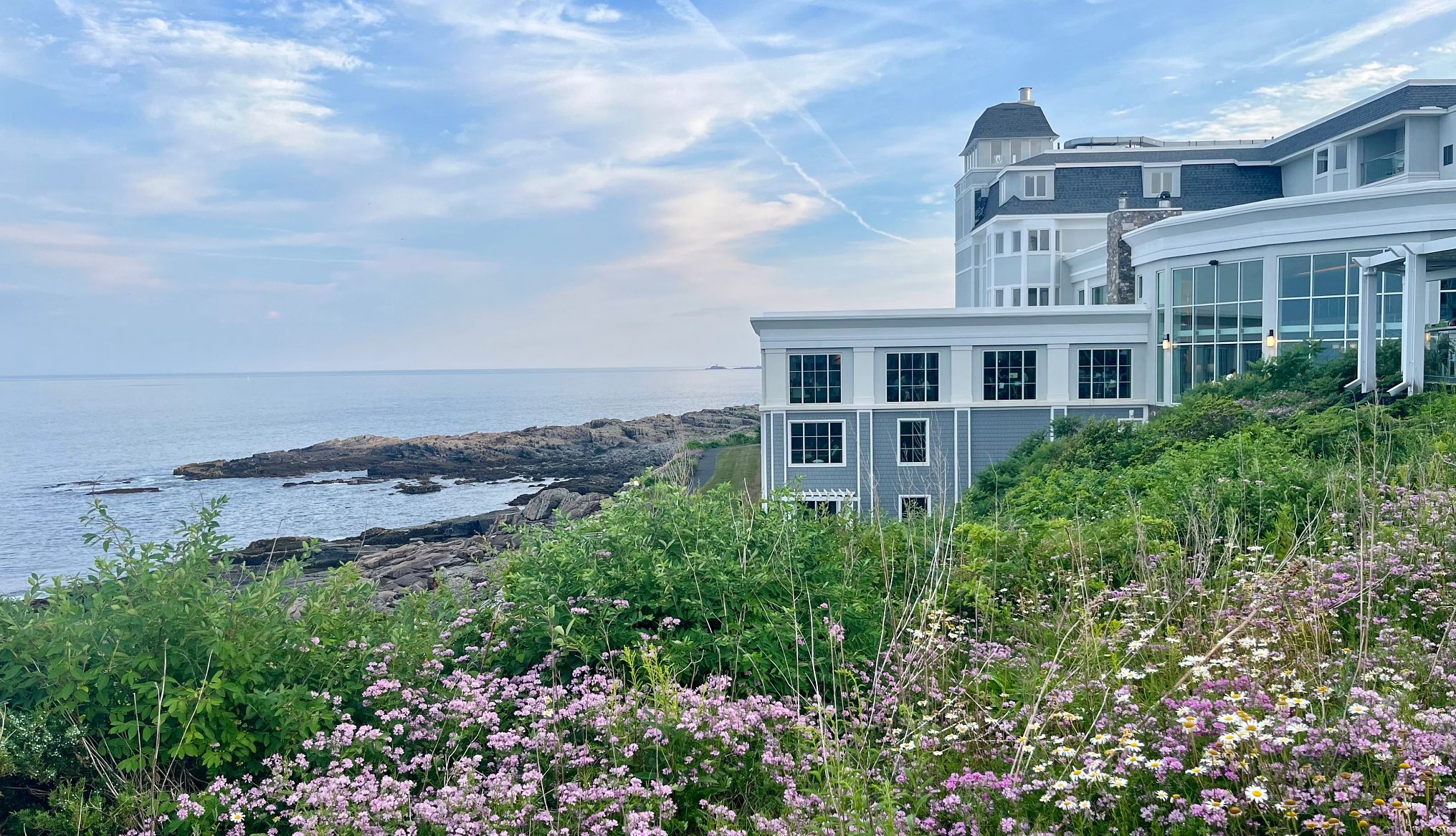 A Writer’s Reflection on Traveler’s Guilt View of Cliff House in Cape Neddick, Maine, surrounded by blooming flowers