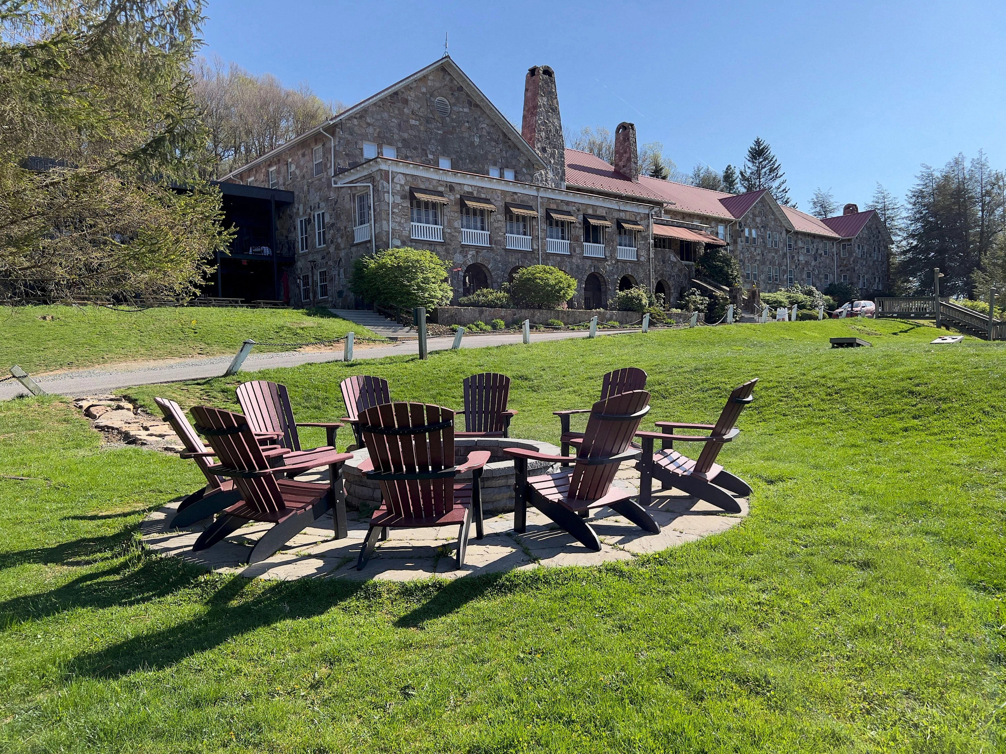 Large stone lodge and retreat property with a circular fire pit surrounded by Adirondack chairs on a green lawn under a clear blue sky