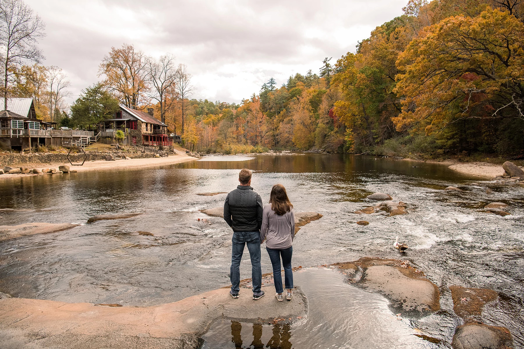 Couple standing on a rocky riverbank overlooking autumn foliage and a retreat lodge in the distance