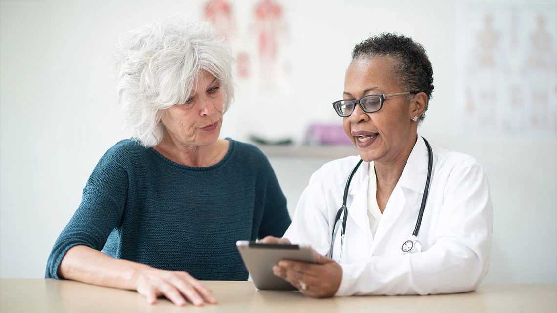 Oak Street Health Primary Care older woman with white hair talking to older female doctor in white coat using a tablet in doctor office setting