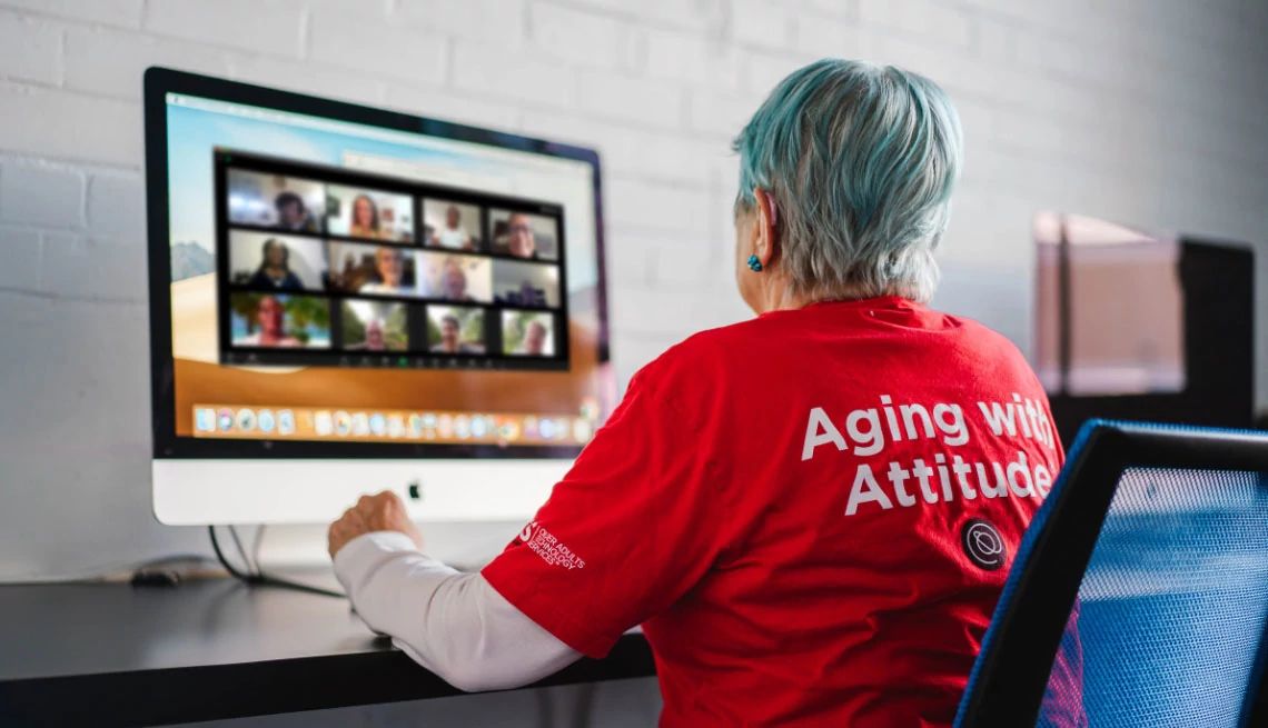 Senior Planet from AARP older woman with back to the camera, working on an mac computer with large screen, woman has short blue colored hair with blue earrings wearing a red t-shirt with Aging with Attitude in white letters on back of shirt