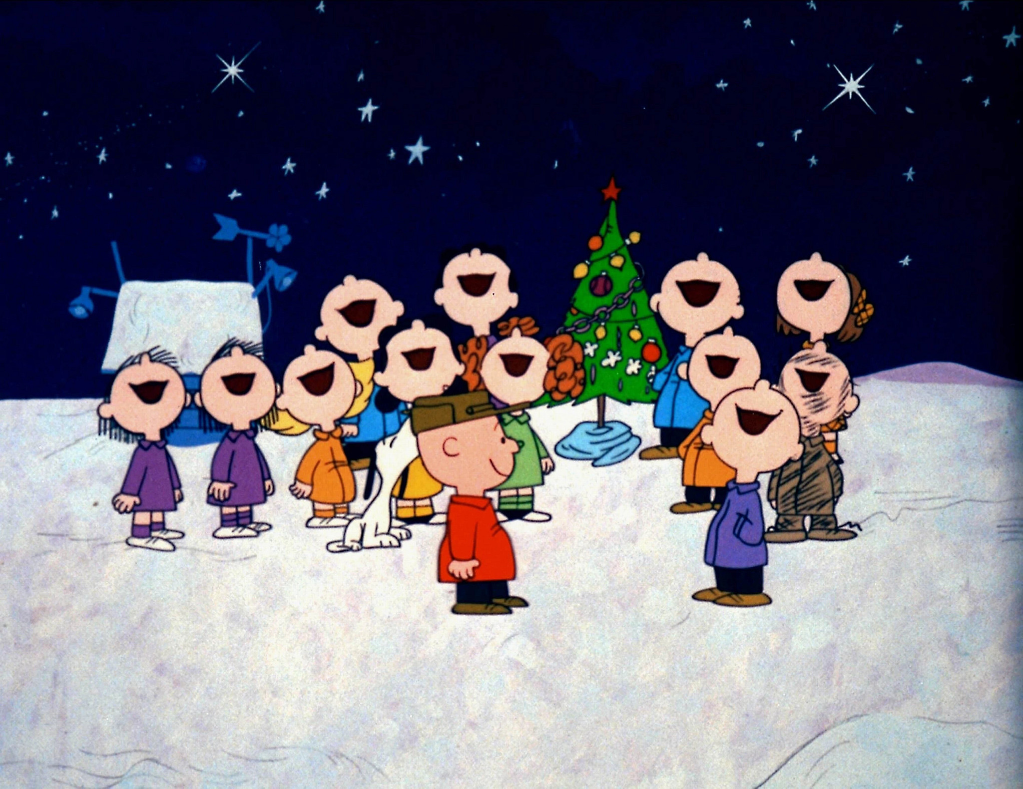 An image from a Charlie brown Christmas is shown. Charlie is smiling while the other Peanuts characters are singing on a snowy, starry night
