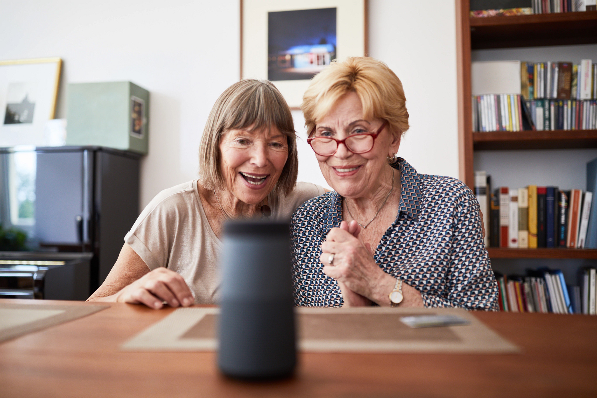 Two senior women looking excited while taking with an interactive voice assistant smart speaker. Excited elderly female friends asking questions to a digital assistant at home.