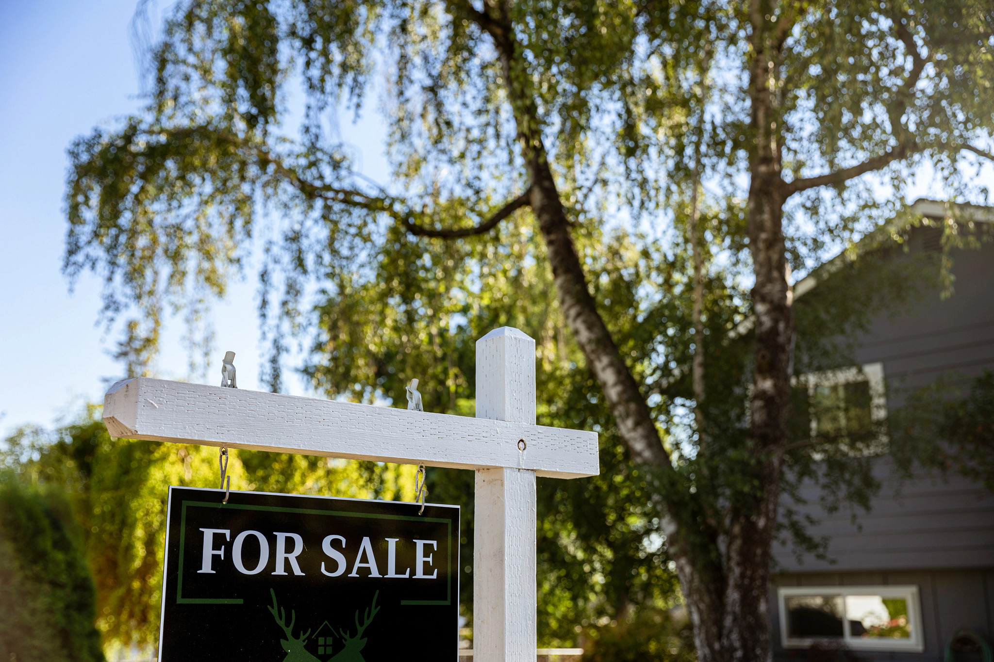 A for sale sign hangs on a wooden post in front of a house.