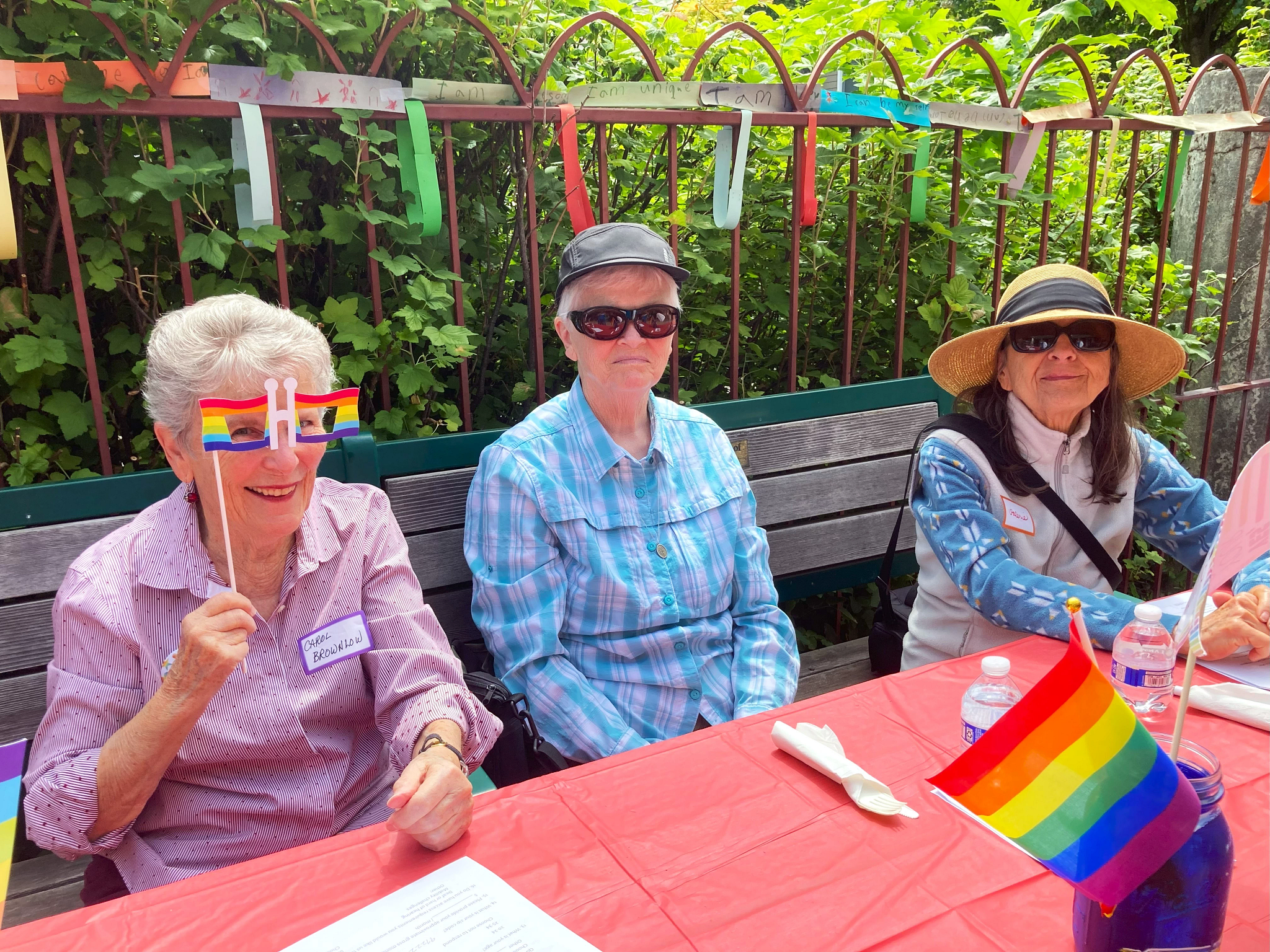 three older adults sit at a picnic table showing off Pride's rainbow colors