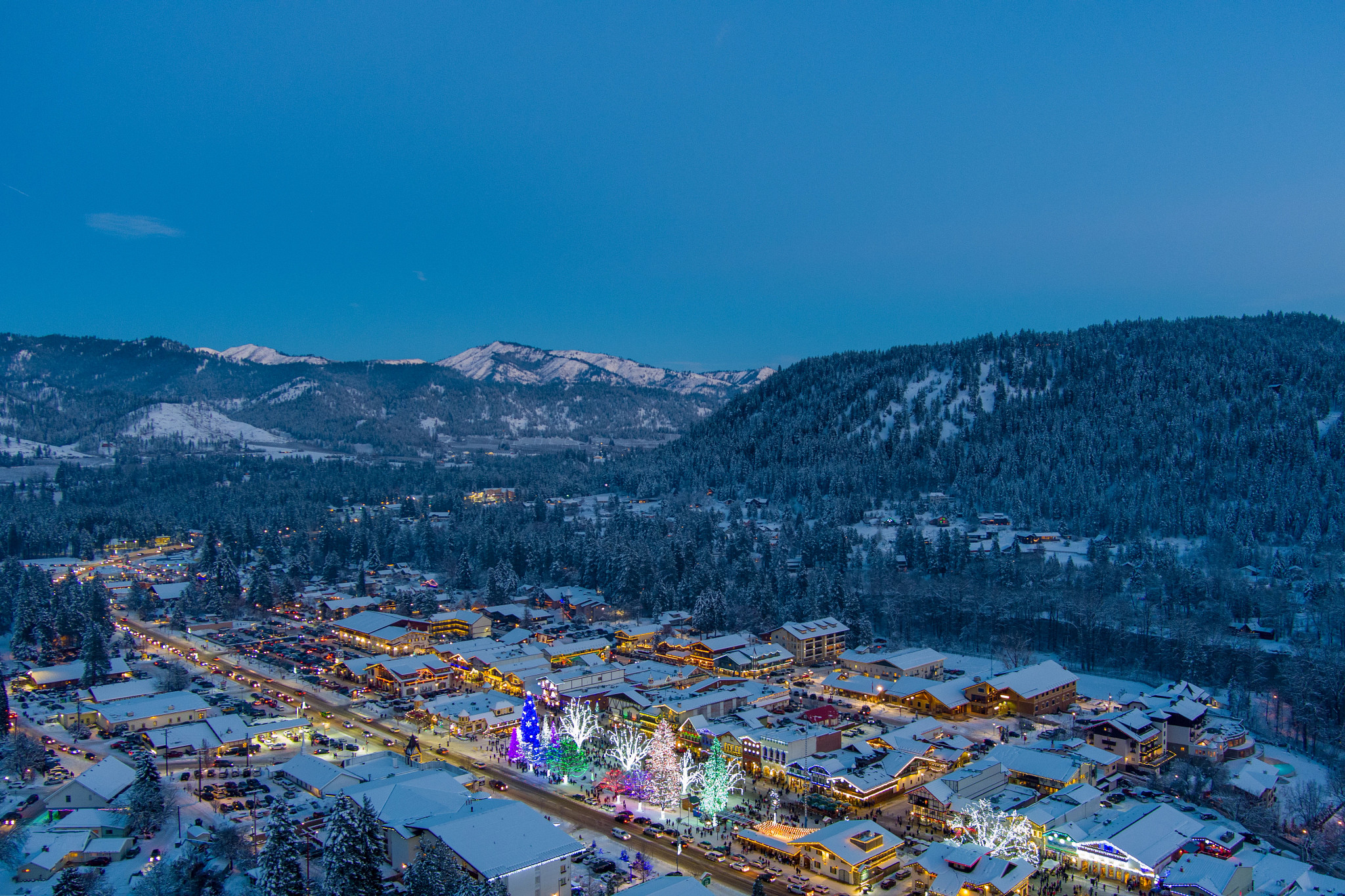 Snow-covered mountain town illuminated with colorful holiday lights at twilight, surrounded by forested hills and snowy peaks.