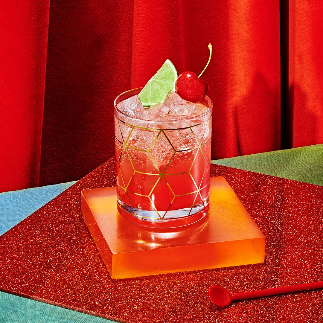 Pink mocktail with ice, lime wedge, and cherry garnish in a geometric glass