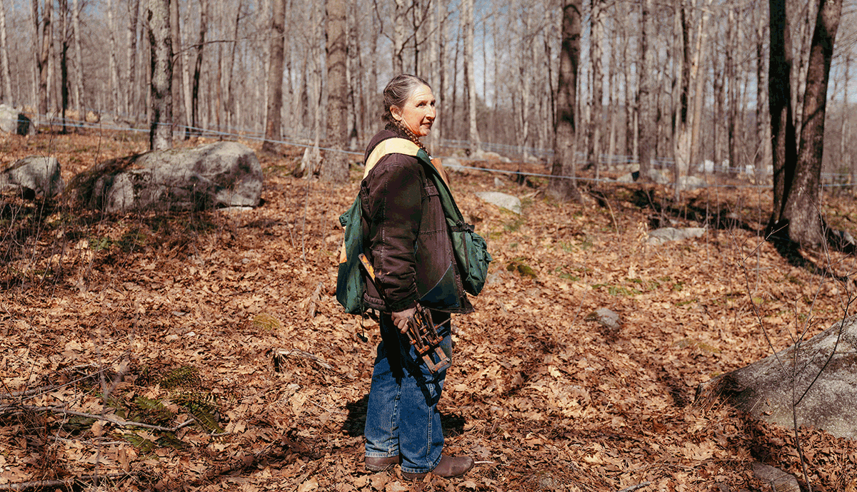 Heidi Bundy walking through a wooded forest with fallen leaves, carrying outdoor equipment and wearing a backpack