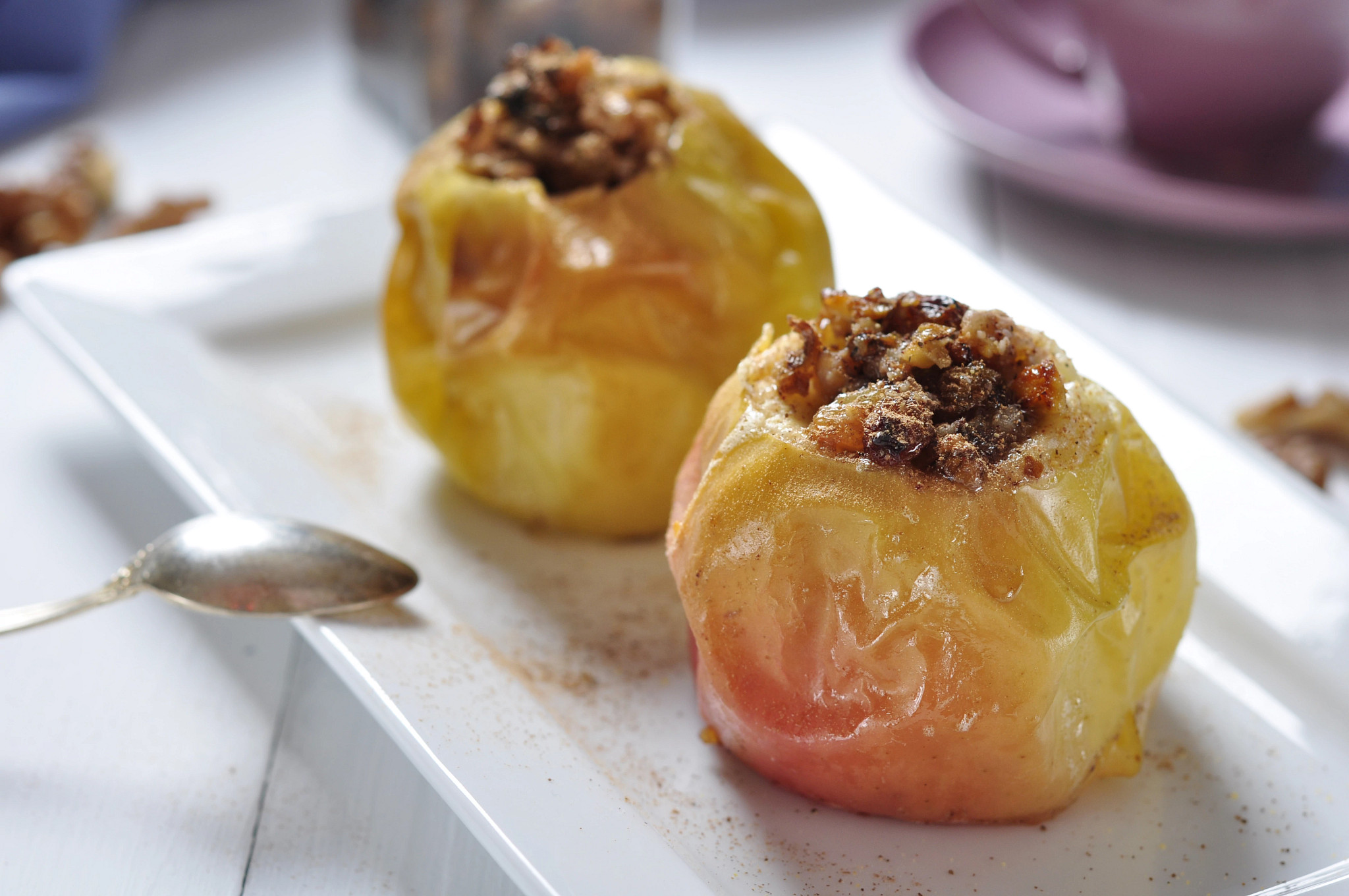 Two baked stuffed apples filled with a cinnamon walnut topping on a white serving platter. A spoon rests nearby