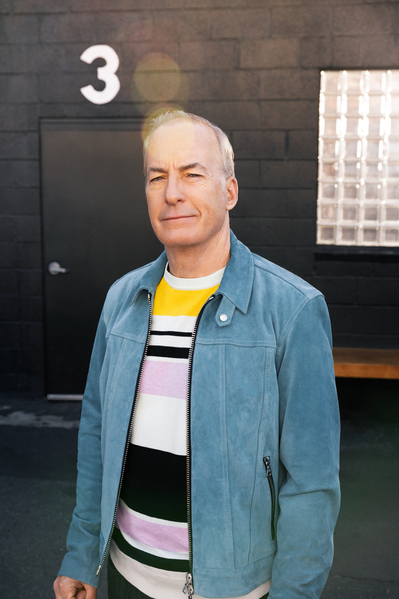 bob odenkirk posing for a portrait in a black-painted studio space, wearing a blue suede jacket and a striped shirt
