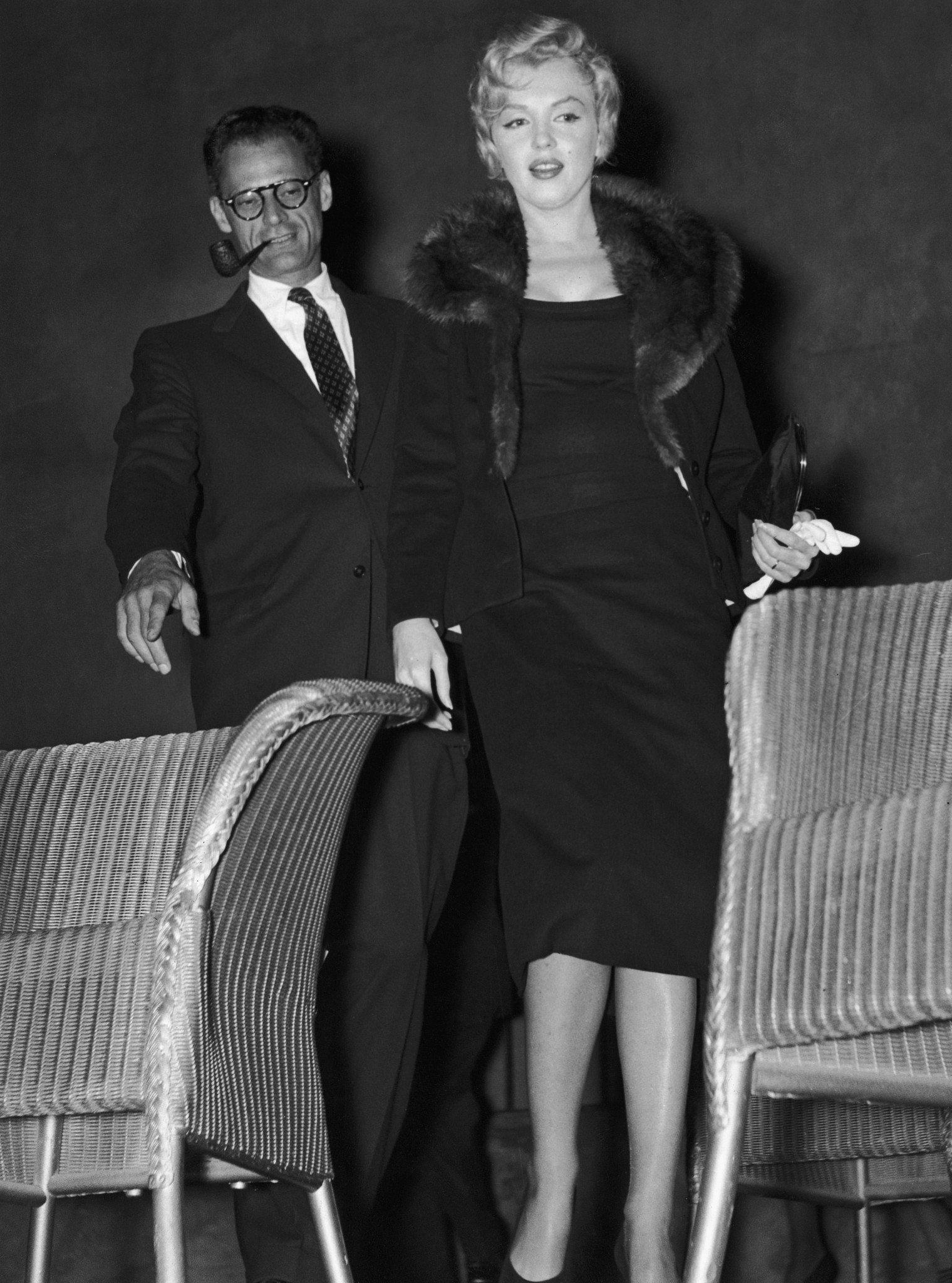 Black-and-white photo of Arthur Miller and Marilyn Monroe dressed in formal evening attire standing indoors near upholstered chairs, with Monroe wearing a dark fitted dress and fur-trimmed coat and holding gloves, and Miller in a suit with glasses and a pipe