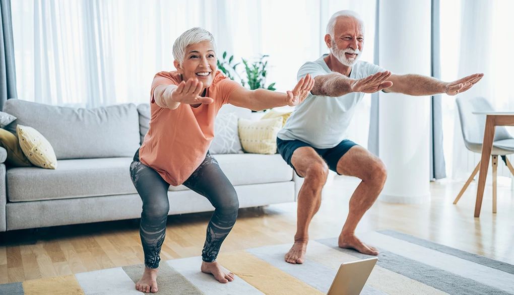 Try exercising during TV commercial breaks A man and woman doing air squats in the living room of a home