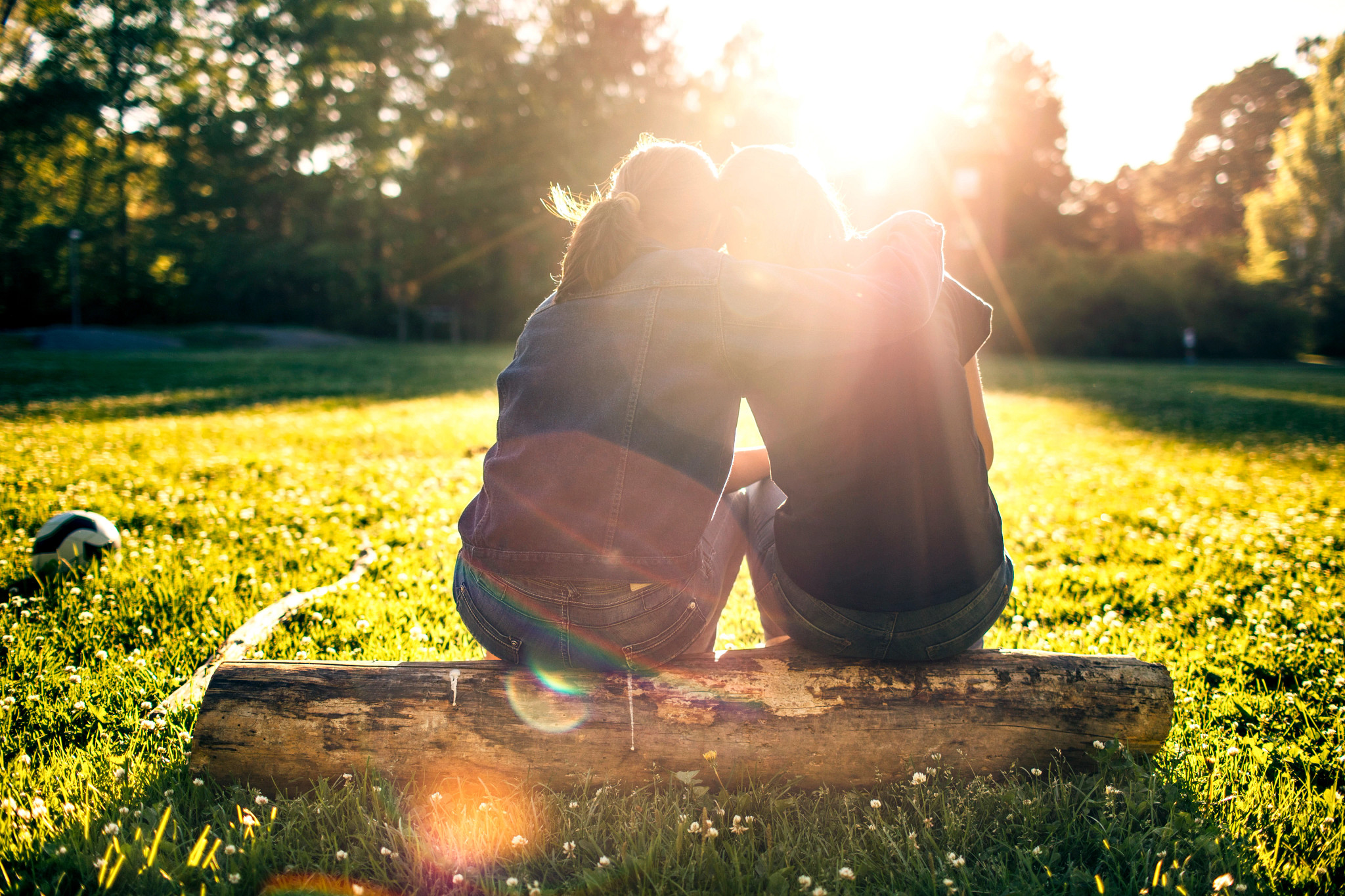 a photo shows two young adults sitting on log, watching the sunset behind a row of trees
