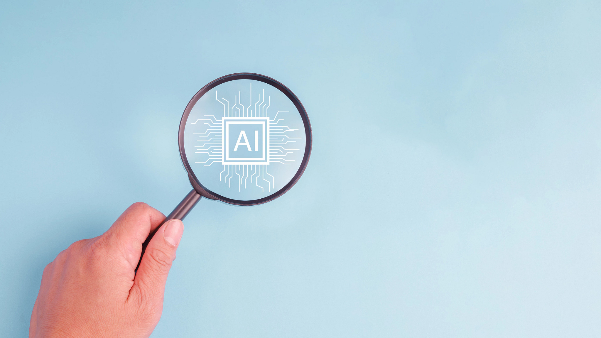 A hand holding a magnifying glass over a pastel blue background with the word AI on chip. Concept of artificial intelligence technology adoption