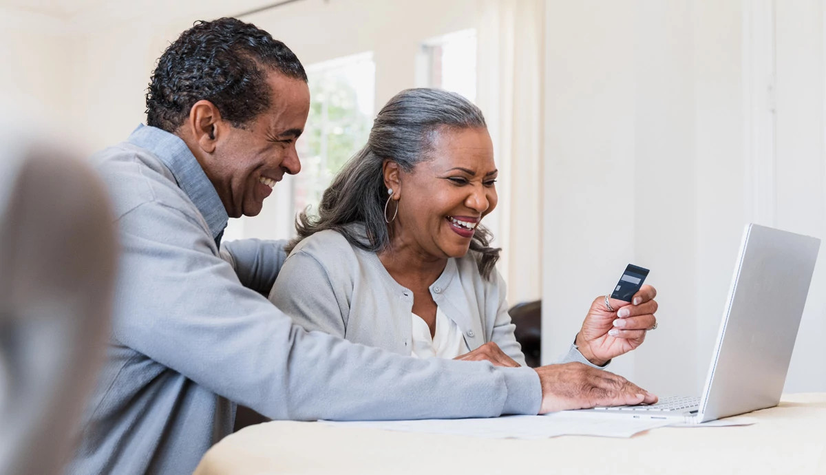 Two older adults smile as they look at a phone and laptop to complete an online task.