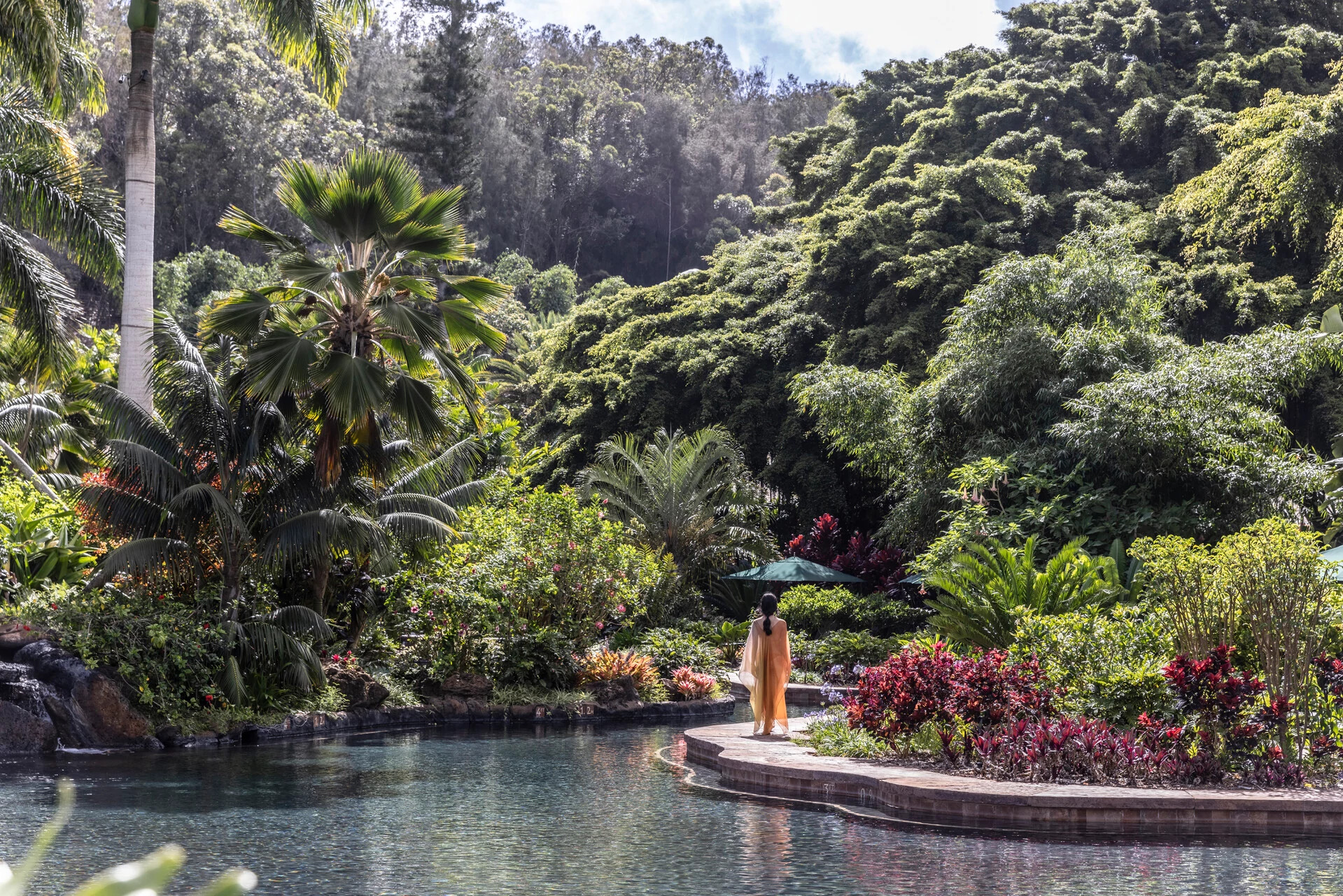 a person walking near a pool surronded by tropical trees and plants