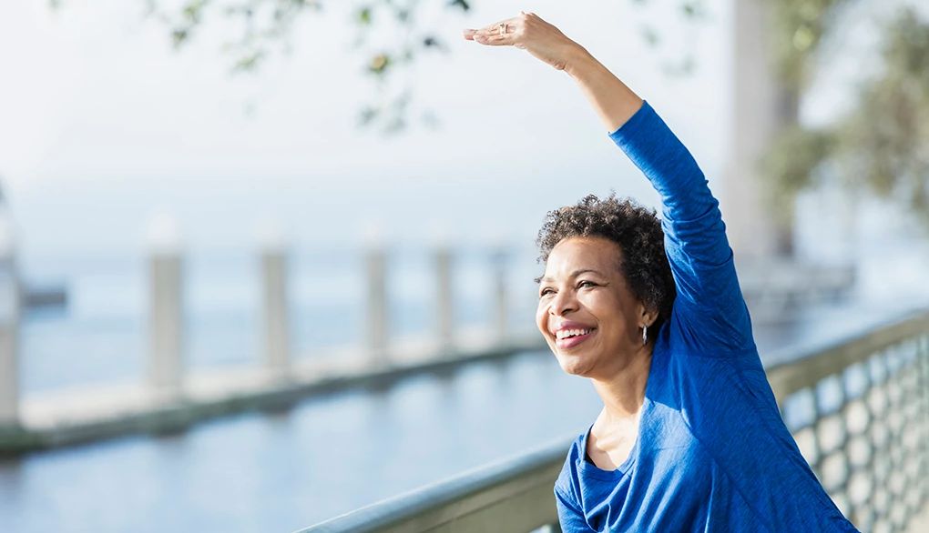 Exercising can make a difference in women's brain health A woman stretching her arm over her head along a waterfront
