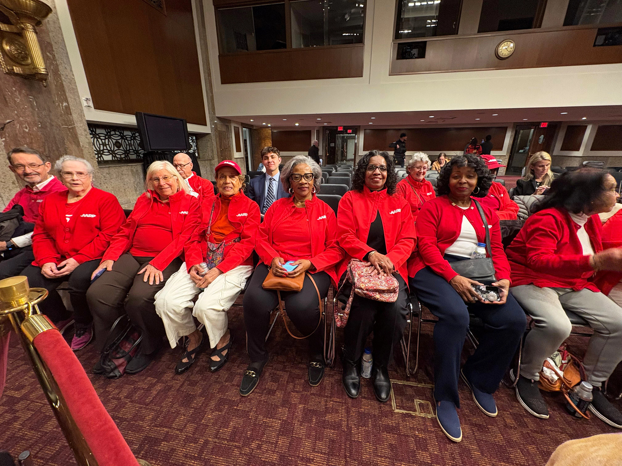 A group of people in red AARP jackets sit in a committee room.