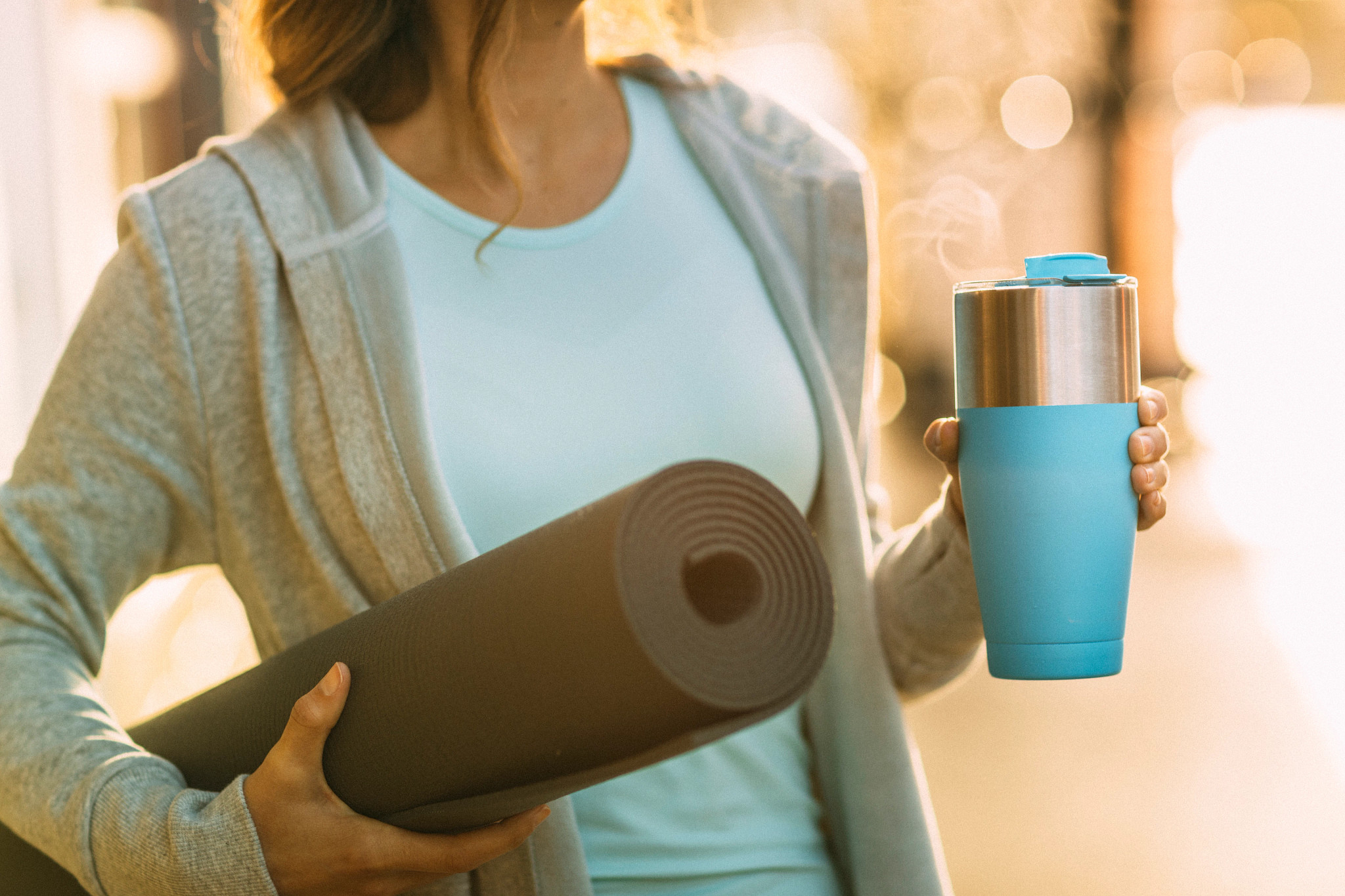 A woman holding a yoga mat in one hand and a coffee mug in the other