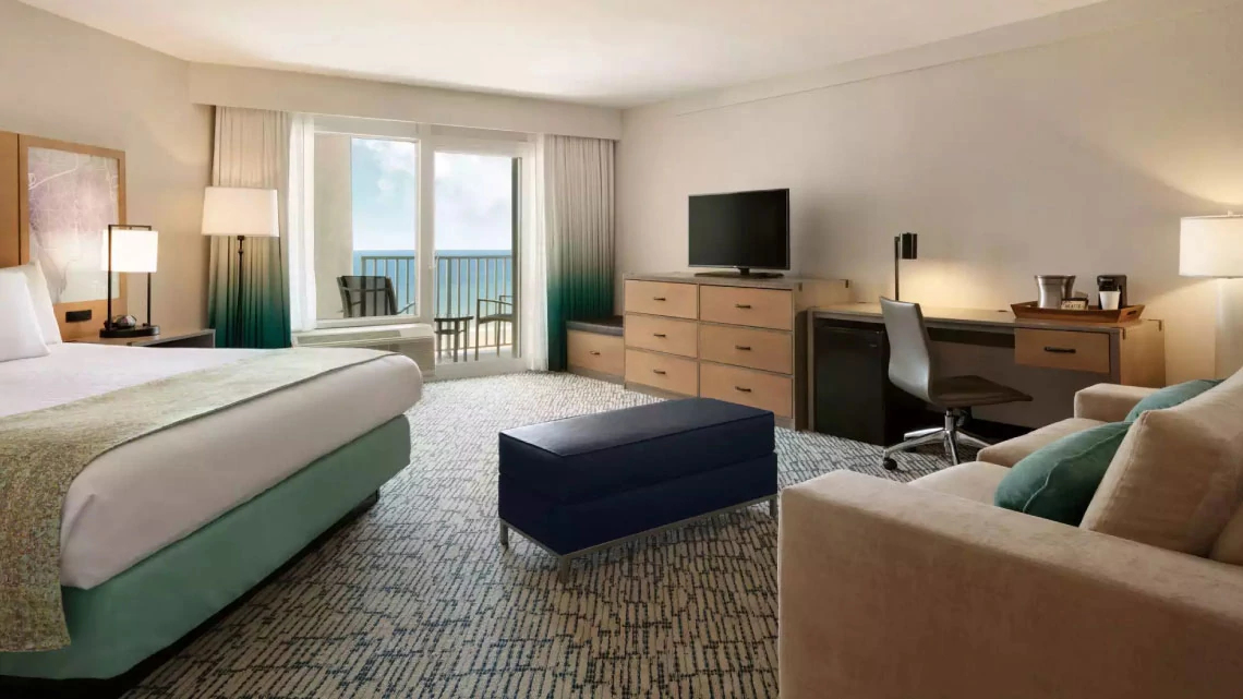 king-size bed in a hotel room with a balcony looking out at the ocean with multiple lamps, desk area, tv, couch, bench