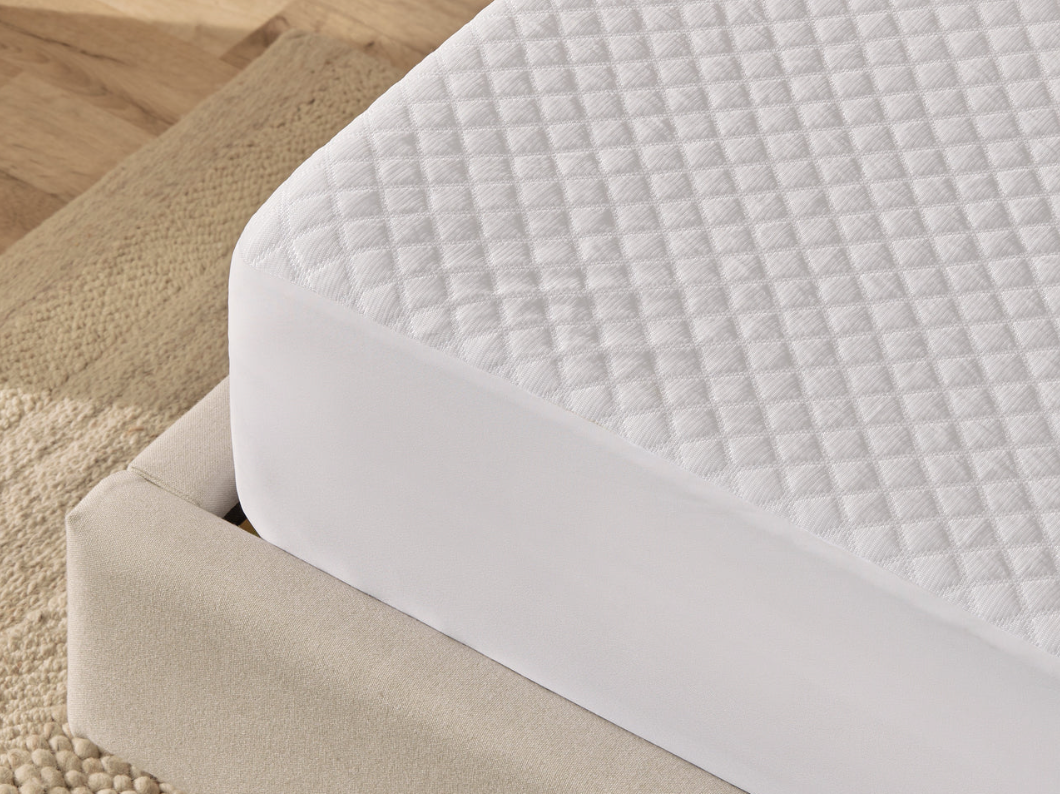 The corner of a mattress on a bed frame with the The Mattress Protector by Silk and Snow on it