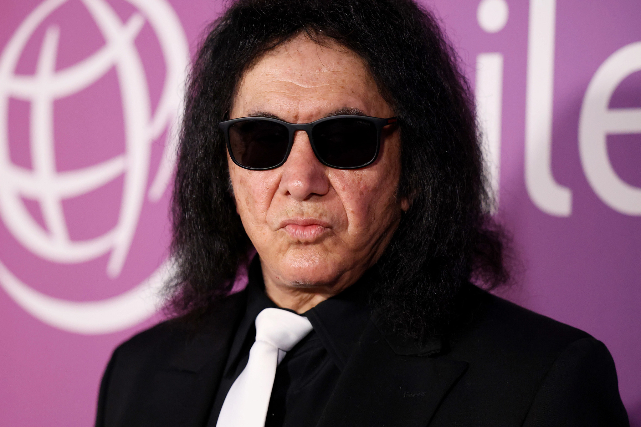 Gene Simmons in a black jacket, white tie and sunglasses