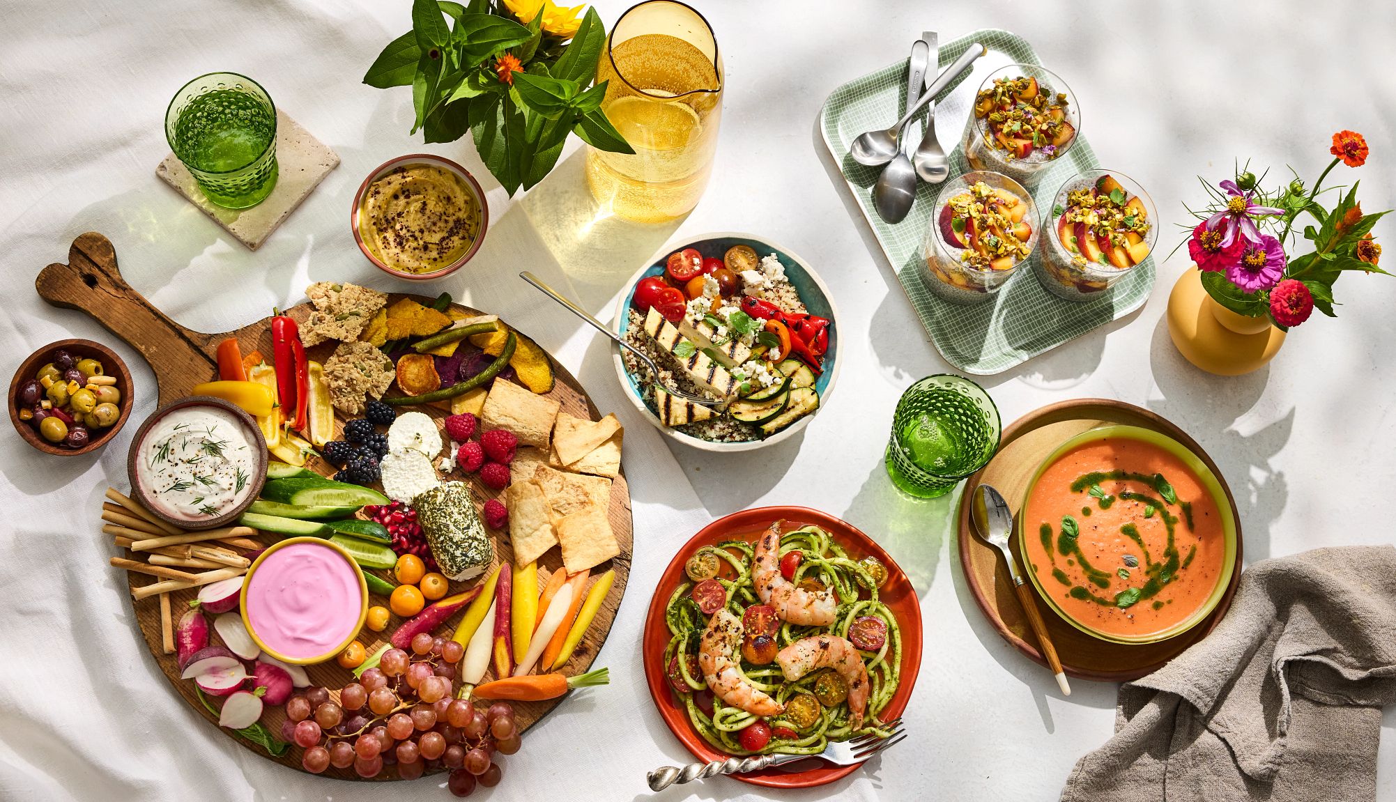 Farmers Market Menu A photo with an overhead view shows a mediterranean grazing board, zucchini spirals with grilled prawns, spicy stone fruit gazpacho, peach chia custard parfait and a grilled vegetable grain bowl. Olives, flowers and ice water are also on the table