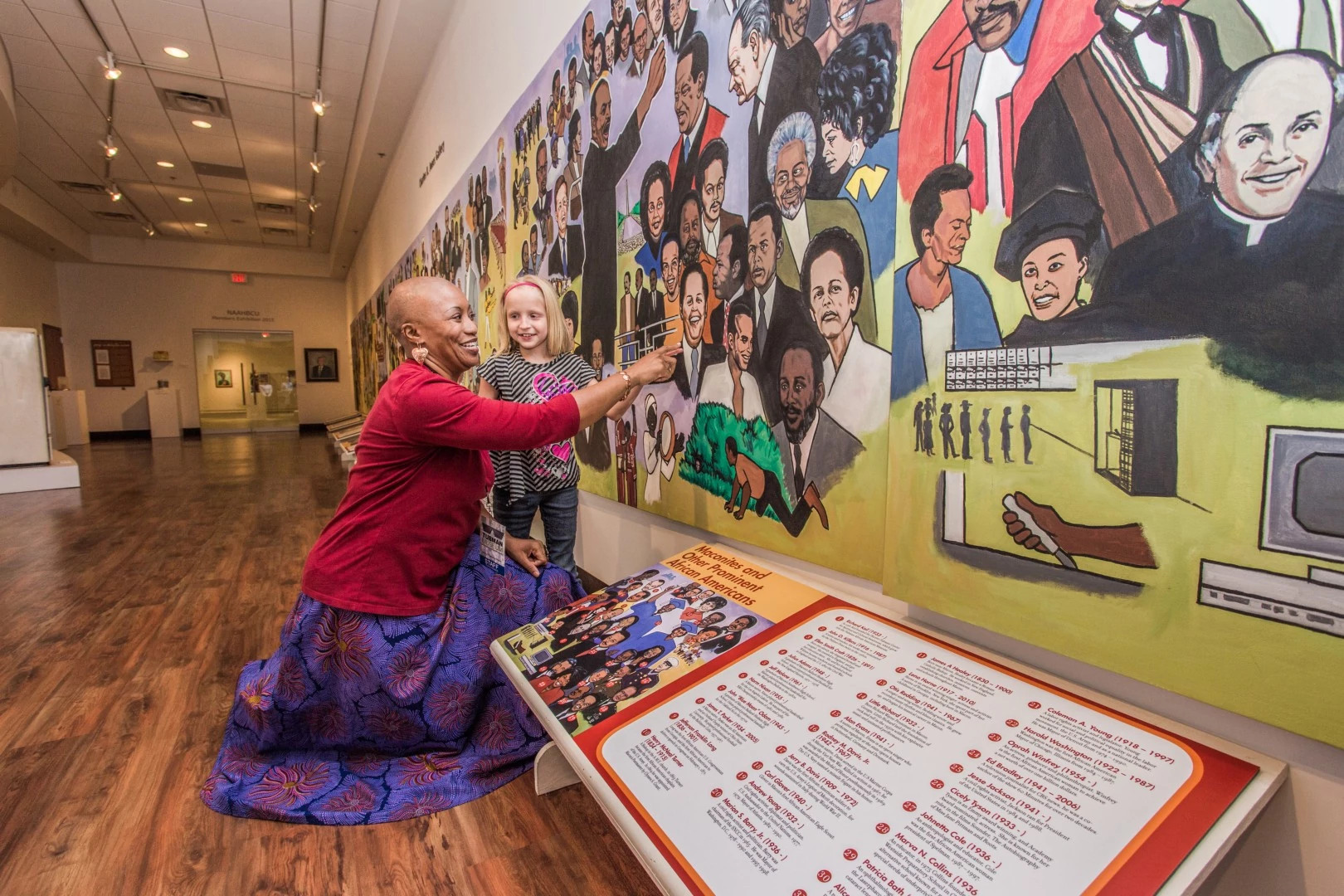 an adult bends down next to a child, pointing out something on a mural 