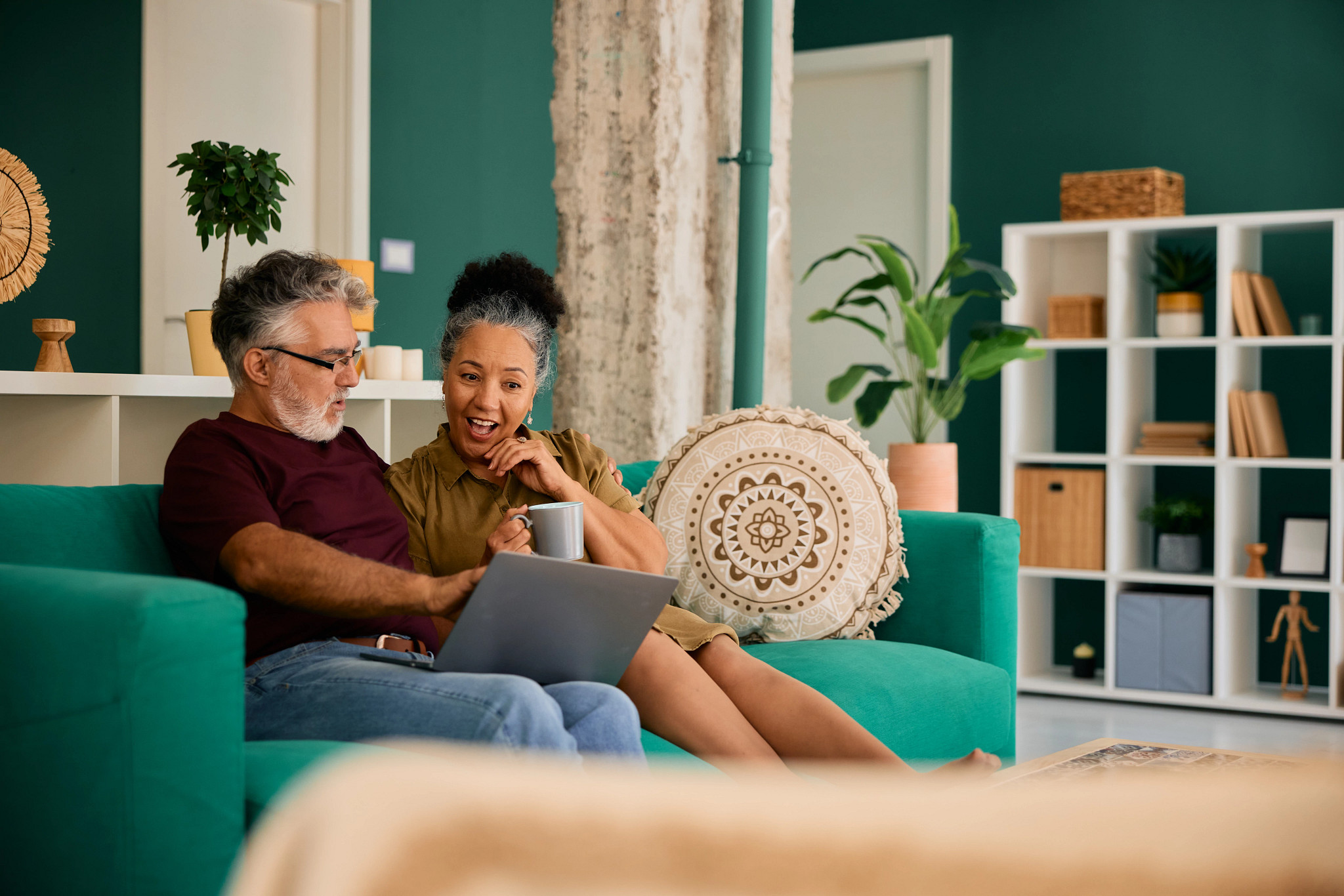 Mature couple sitting on a cozy sofa at home, looking surprised at their laptop screen, with the woman happily holding a steaming cup of coffee, enjoying their time together