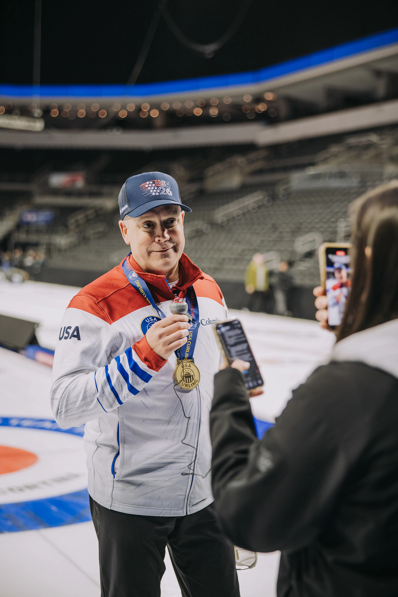 A smiling Rich Ruohonen wearing a Team USA jacket and a blue "USA 26" baseball cap while being interviewed on the ice; he is wearing a gold medal around his neck