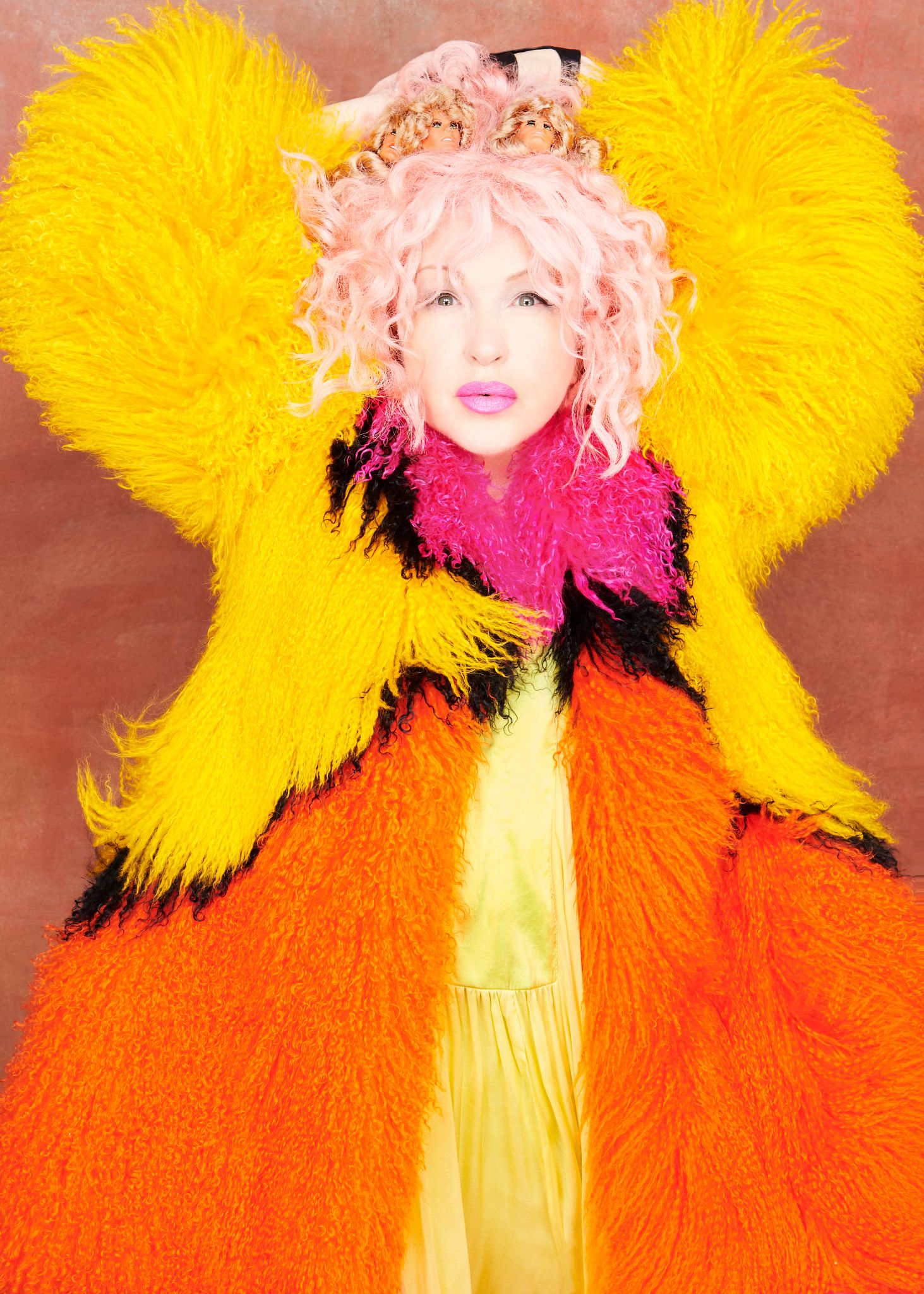 Singer Cyndi Lauper wearing a colorful yellow, red and pink outfit that covers her arms along with three toy doll heads attached to her hair